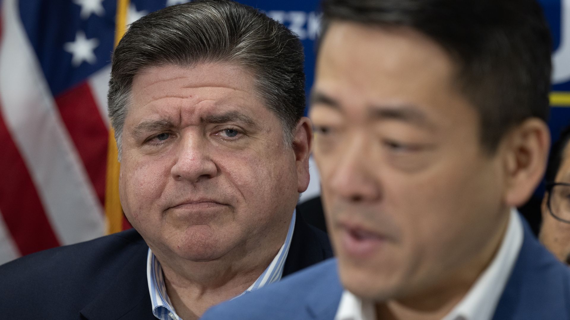 Two men in suits at a press event. The man in the foreground is out of focus and speaking. The serious man in the background has gray-streaked hair, a blue striped shirt, and an American flag behind him.
