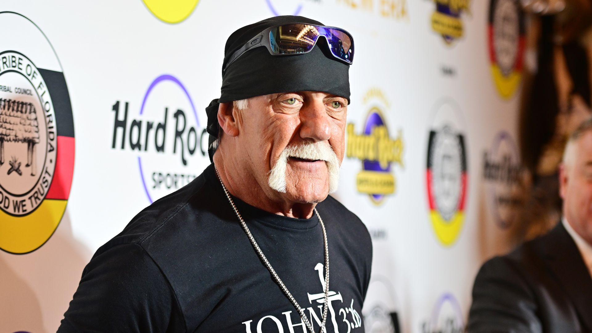 A man with a white handlebar mustache in a black T-shirt with a Black bandana and sunglasses on his head stands in front of a backdrop that reads "Hard Rock sports."