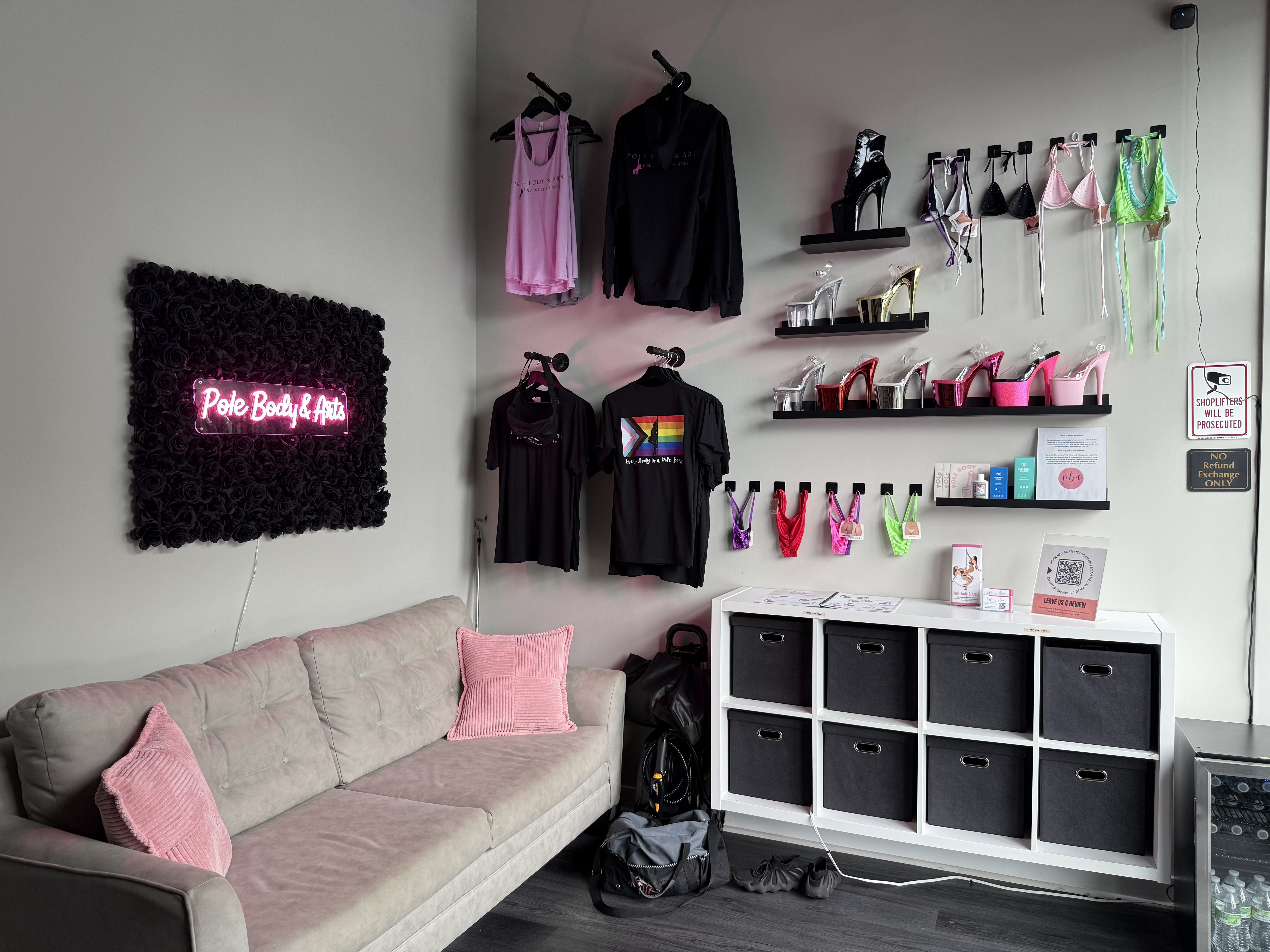 Boutique corner with beige sofa, pink pillows, black rose wall art with pink neon "Pole Body & Arts" sign, apparel, high heels, colorful bikinis, and black storage bins.