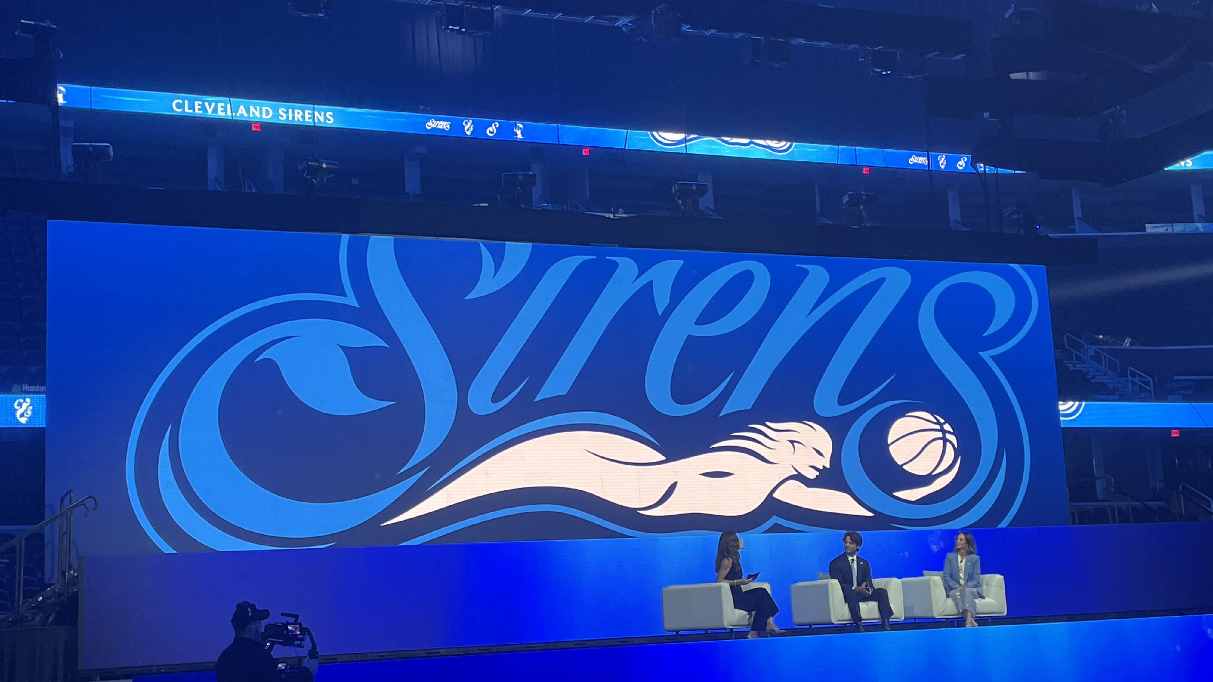 Inside a large arena, a blue-lit stage features a screen with a stylized blue lion and swirls; above it, a blue banner reads "CLEVELAND SIRENS" as three panelists sit on white chairs.