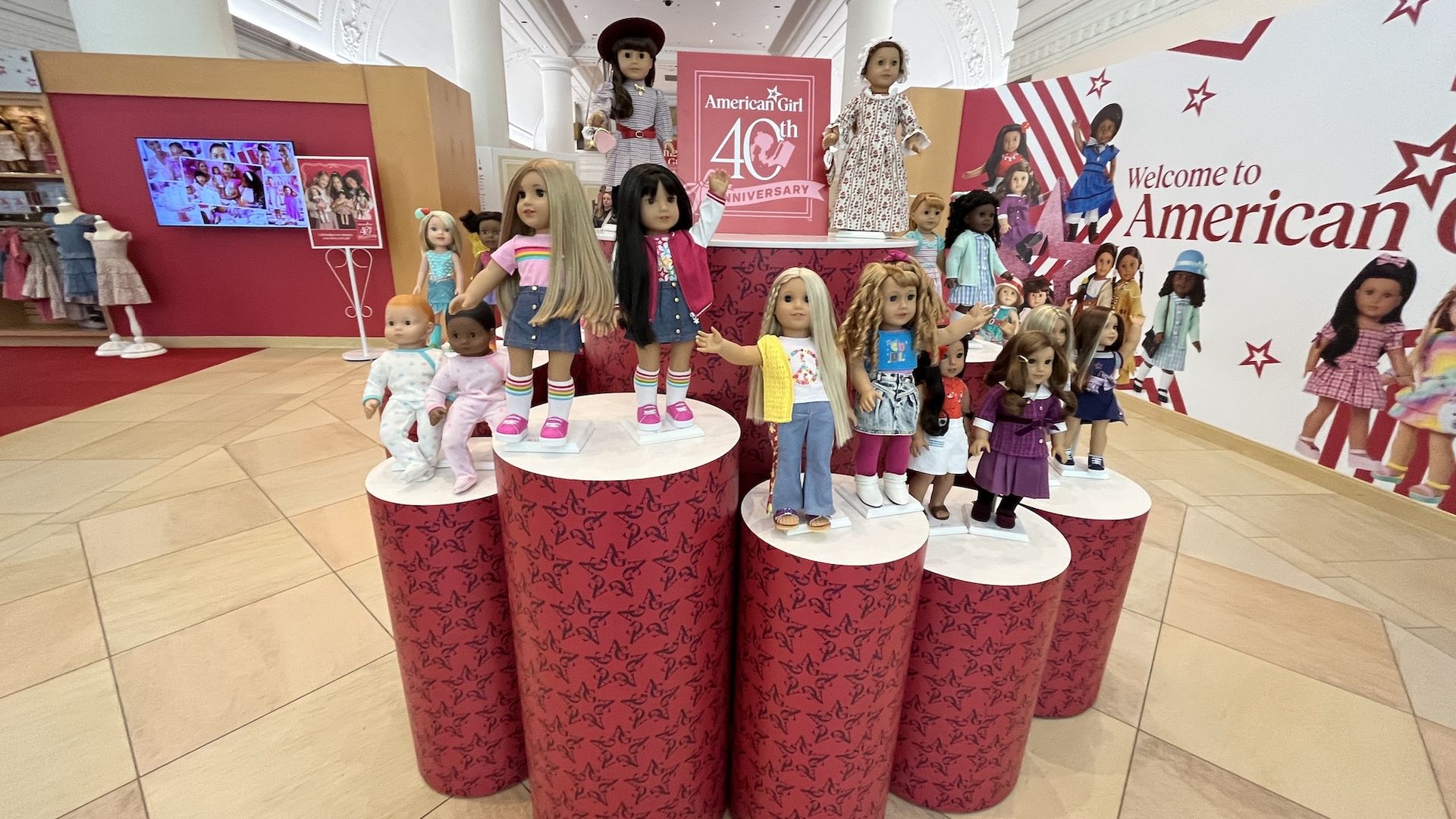 An American Girl store display of numerous dolls posed on red patterned cylinder pedestals, with a pink 40th Anniversary panel and starry wall graphics in a bright, arch-lit showroom.
