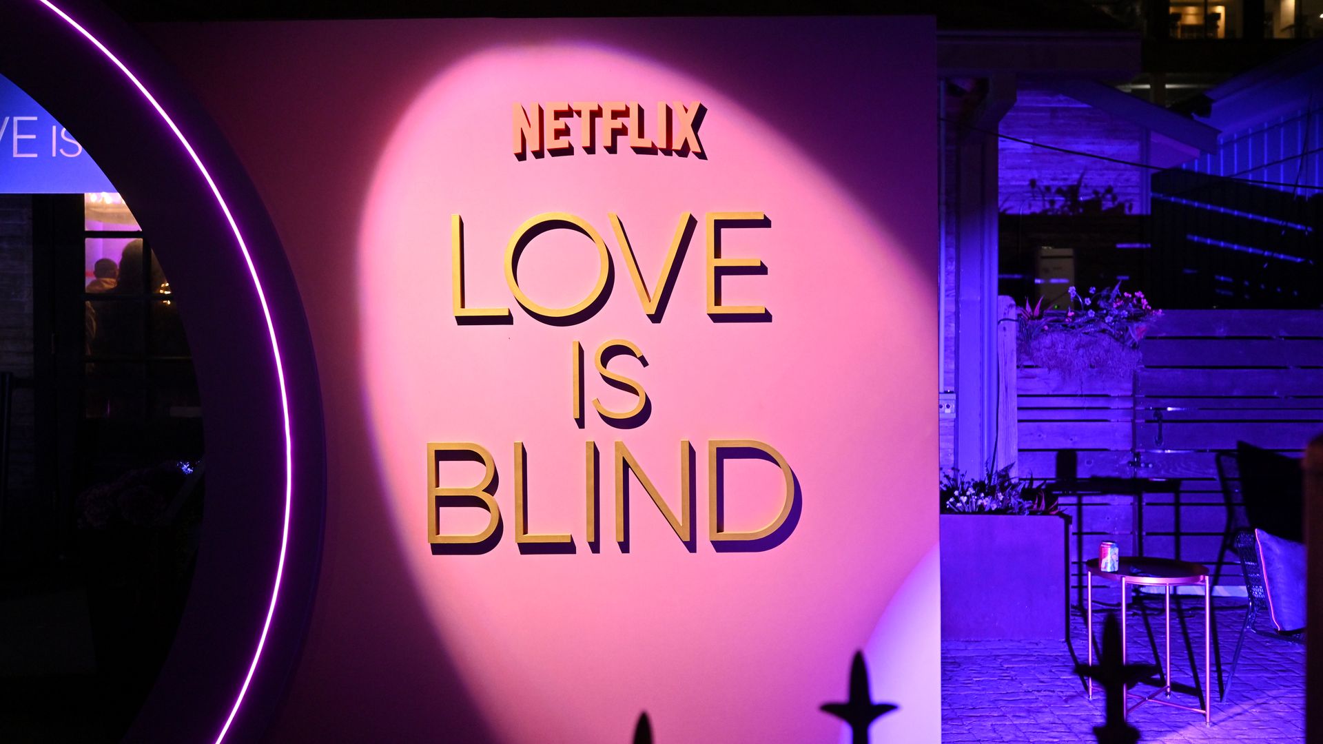 Pink wall lit with a spotlight, featuring yellow letters spelling "NETFLIX LOVE IS BLIND" in a nighttime outdoor setting with purple lighting accents.