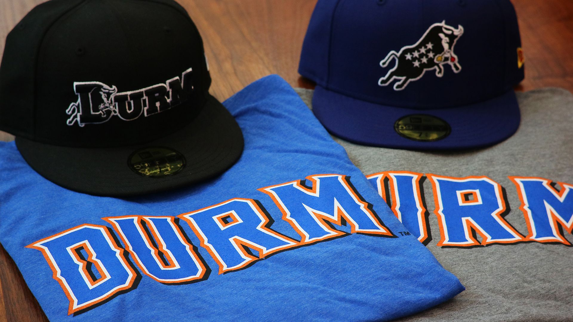 Two baseball caps rest on folded shirts on a wooden surface: a black cap with white embroidery and a royal blue cap with a white longhorn logo, beside a large blue T-shirt with orange lettering.