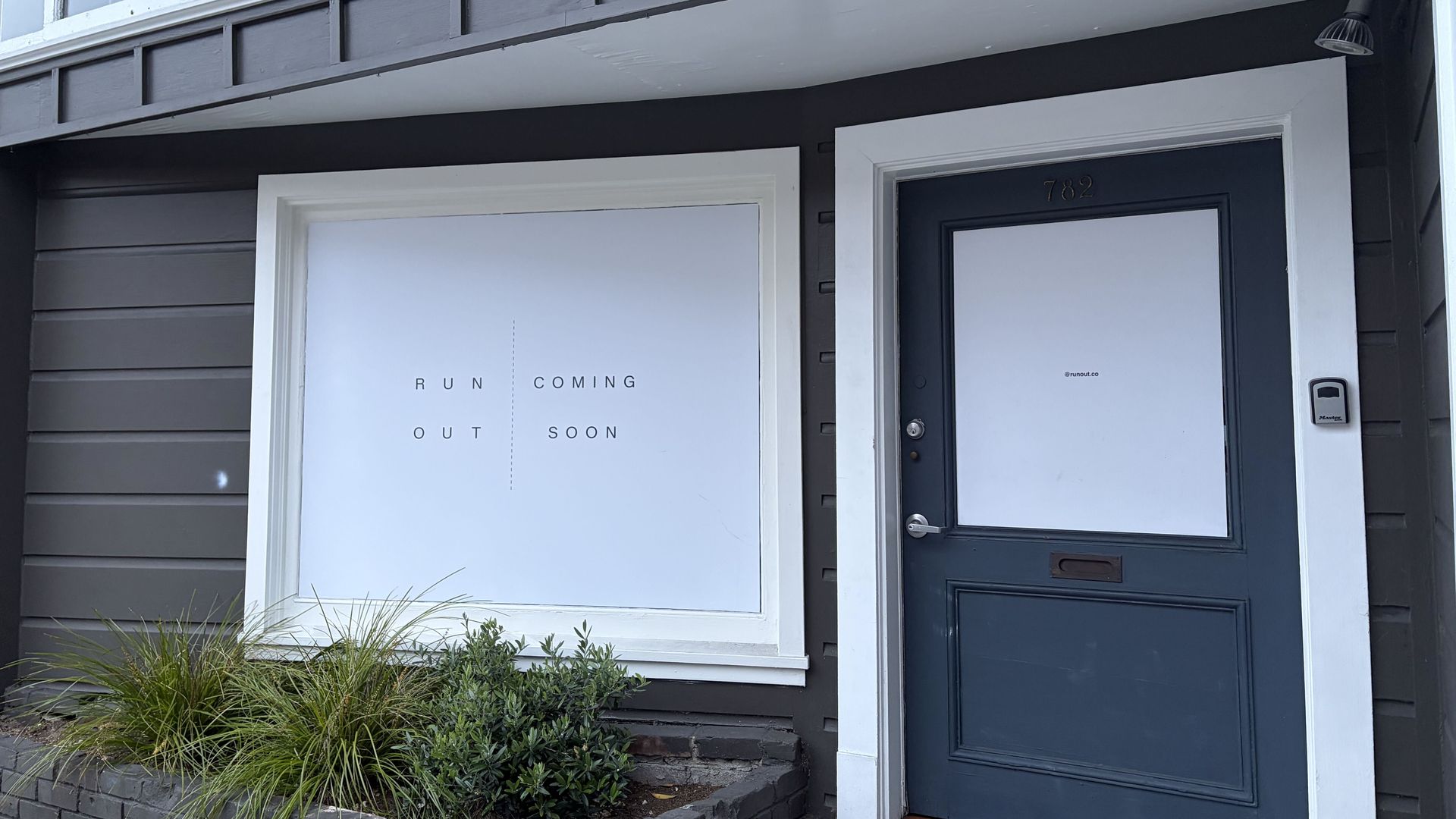 Storefront with a dark gray door and white trim, a large left window displaying "RUN COMING OUT SOON," gray siding, a small security device by the door, and a brick planter with grasses.