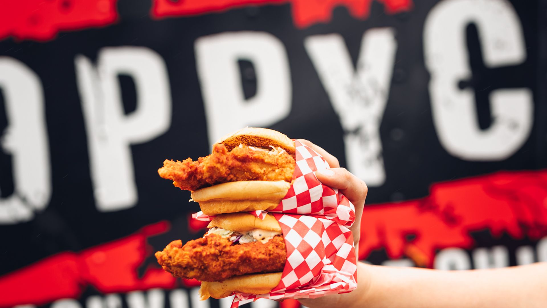 Hand holding two stacked fried chicken sandwiches wrapped in red and white checkered paper, with a black and red background featuring partially visible bold white letters.