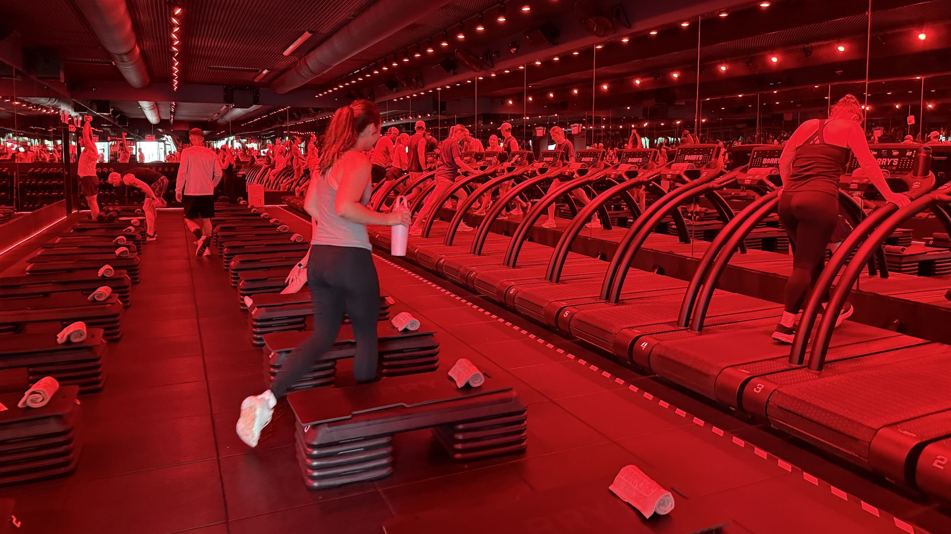 An indoor gym bathed in red lighting, with rows of treadmills and people exercising. Mirrors line the walls, towels scattered on the steps, exposed ceiling ducts and red spotlights.