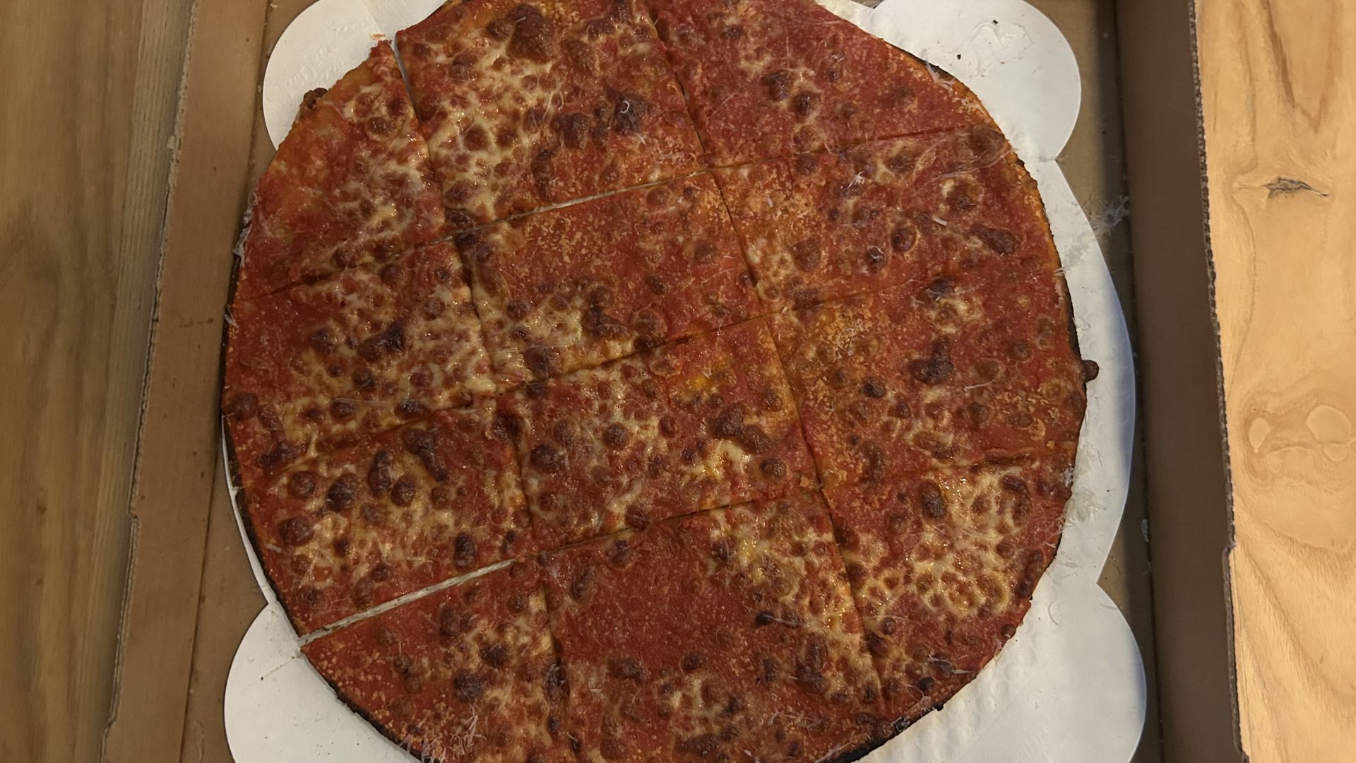 A large, thin-crust cheese pizza with a dark, crispy edge, cut into square pieces, placed on a white paper round inside a cardboard pizza box on a wooden surface.