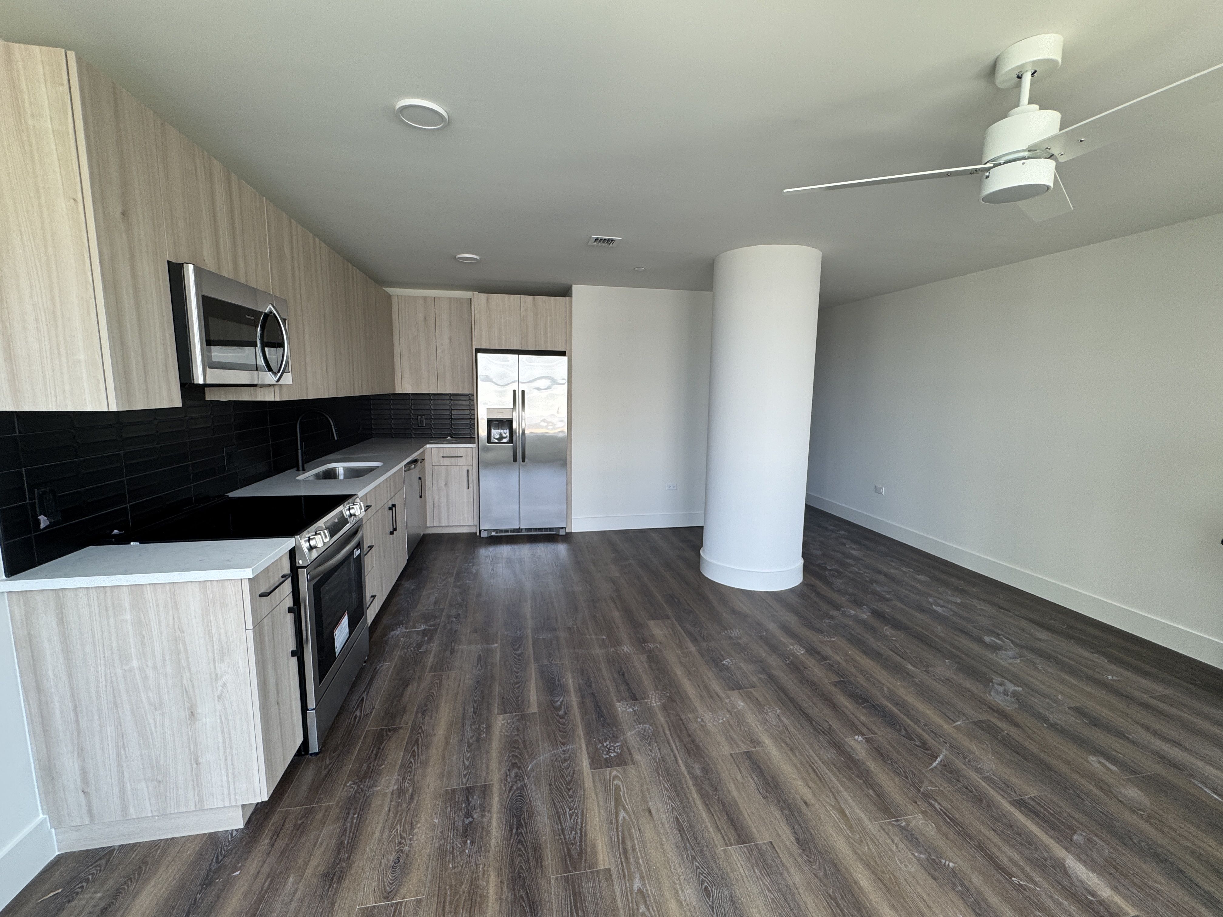 Modern kitchen with light wood cabinets, black tile backsplash, stainless fridge and microwave, and range. White ceiling fan overhead. Large white cylindrical pillar near center; dark wood floors.