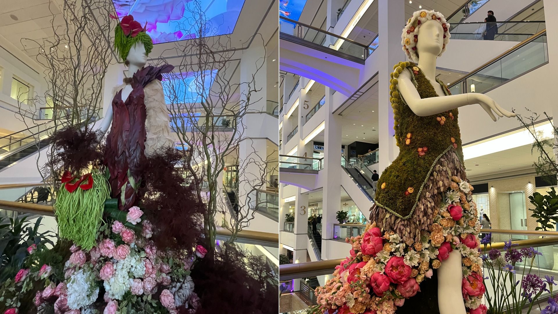 Two fashion mannequins in a mall atrium. Left: white mannequin in a burgundy-and-white gown among bare branches and pink flowers. Right: moss-covered gown with a floral headdress and blossoms.