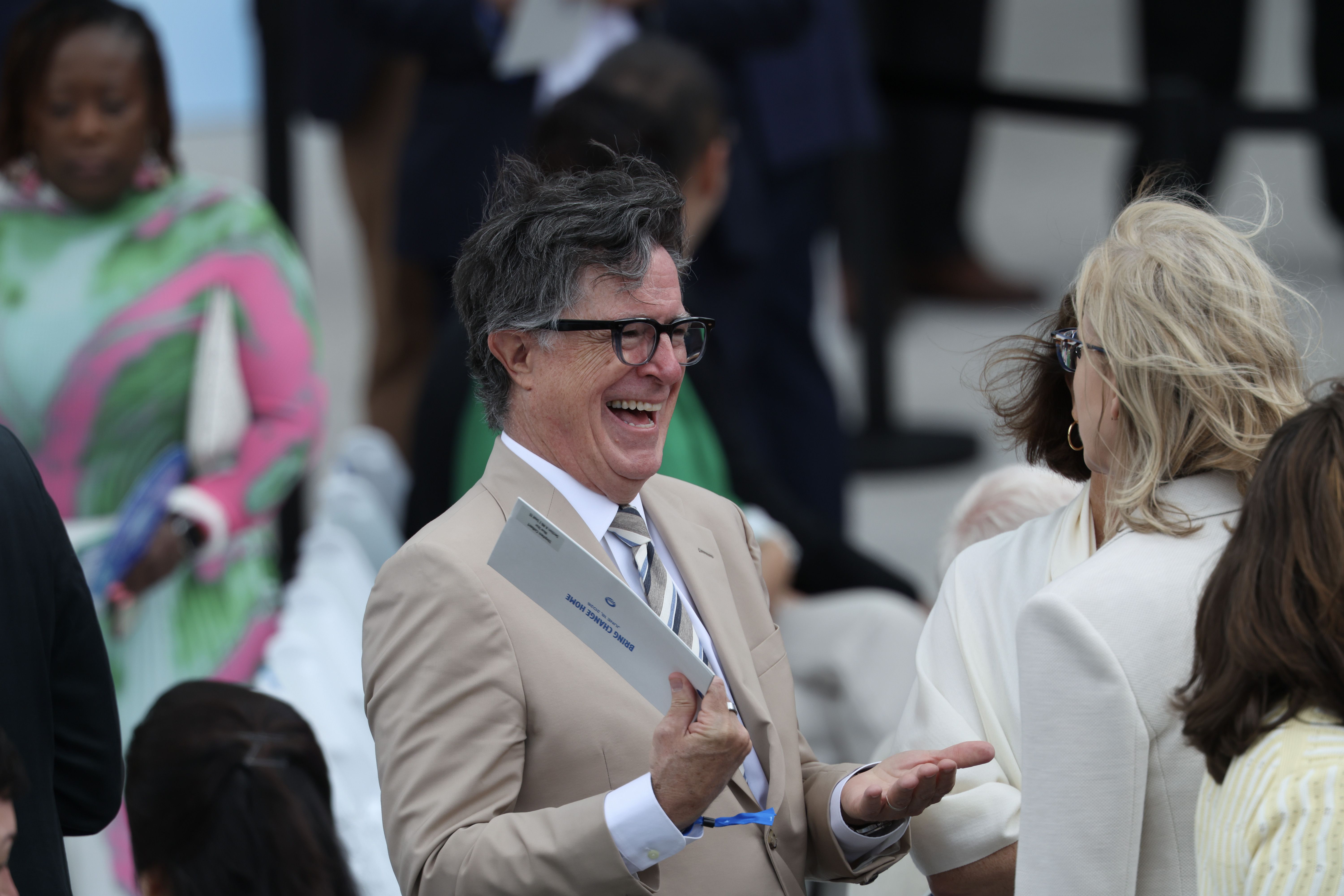 Smiling man with gray hair and glasses in a tan suit holds a program, talking with a blonde woman in white; blurred crowd in the background.