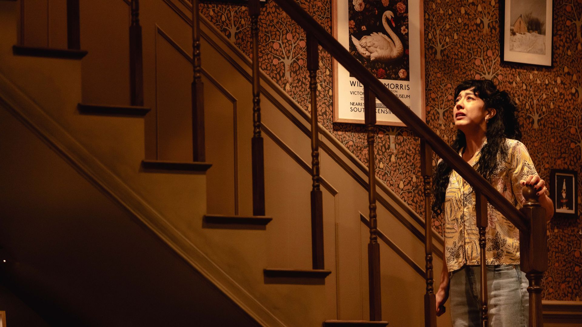 A woman with dark curly hair stands by a wooden staircase, looking up. The wall behind her has orange and brown floral wallpaper with framed pictures, including a swan poster.