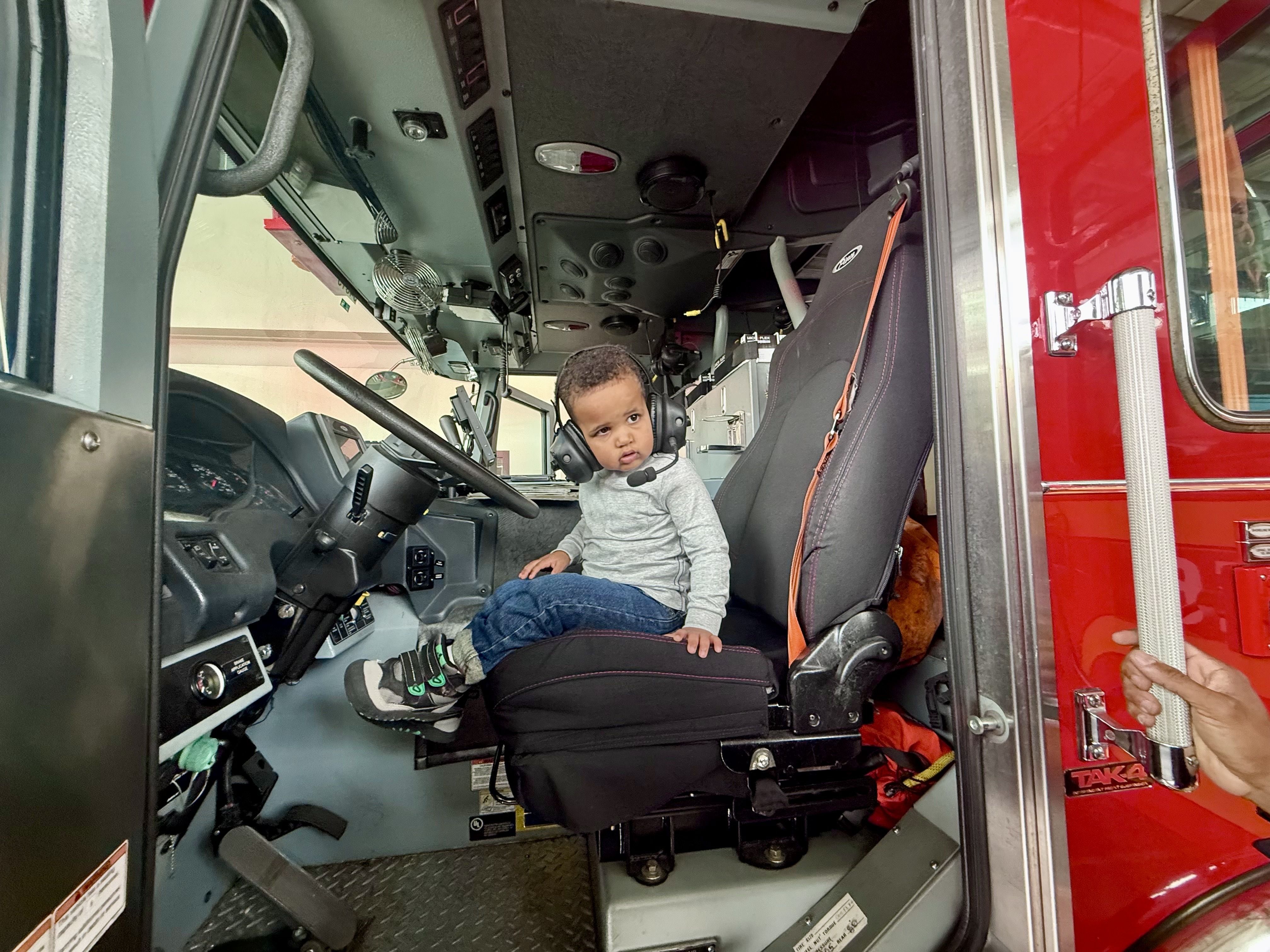 Child wearing a headset sits in the driver's seat of a red fire truck, surrounded by cockpit controls, gauges, and a large steering wheel inside the cab.