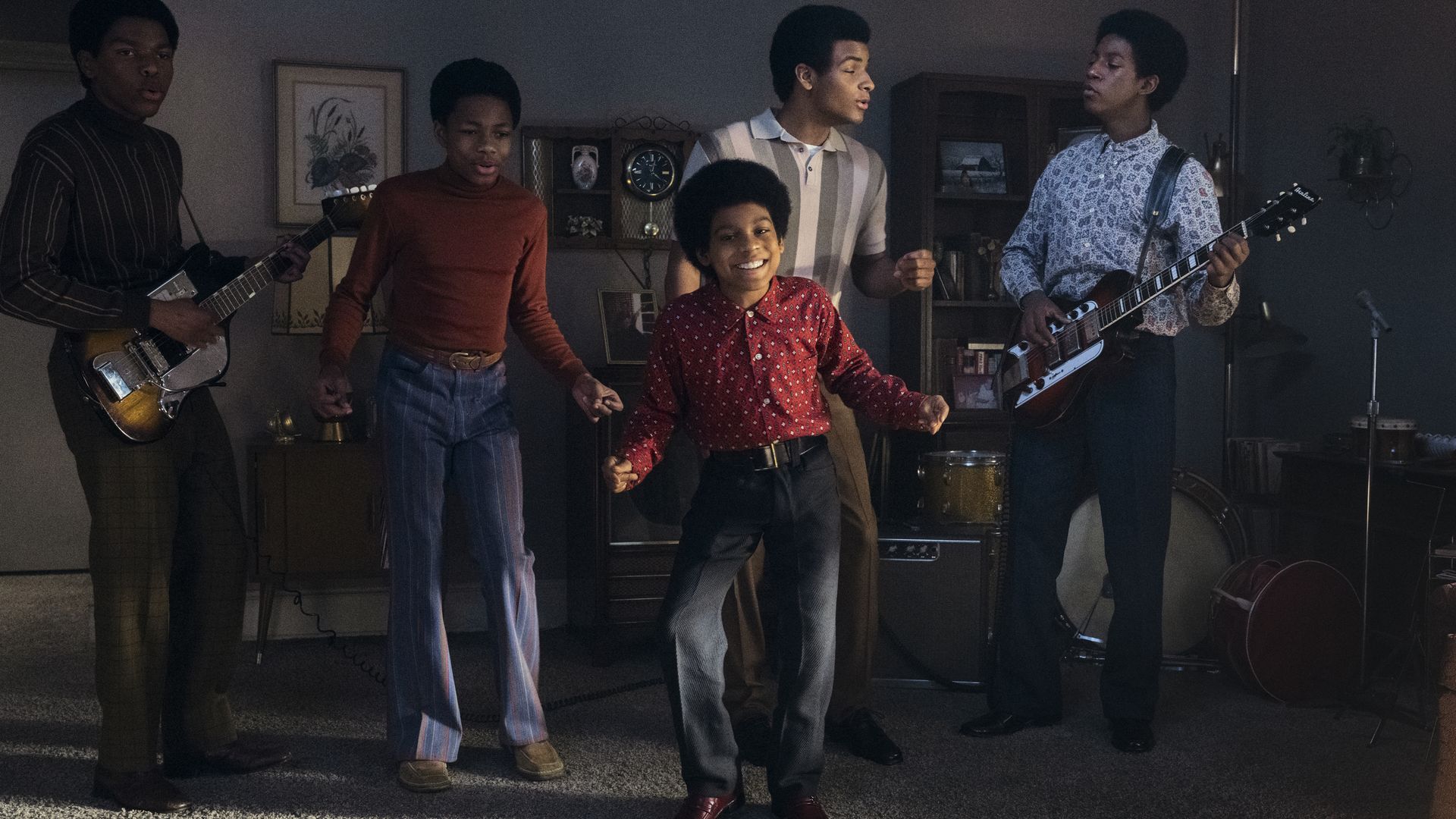 Young men men and a boy form a band in a living room; two guitarists at the ends, center dancer in a red shirt, others sing.