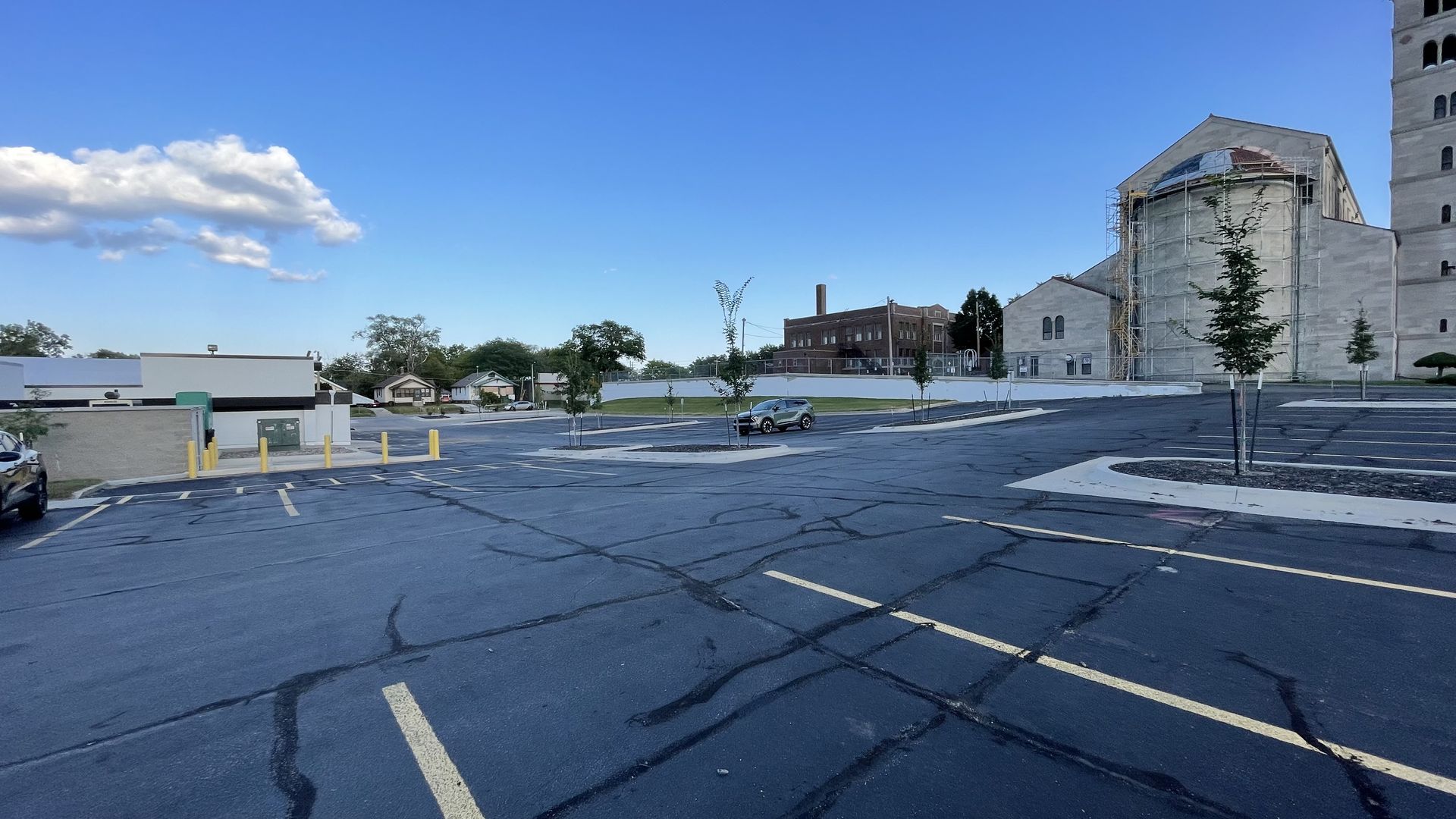 Large mostly empty asphalt parking lot with yellow lines, a few small trees, a silver car, and buildings including one under scaffolding against a clear blue sky.