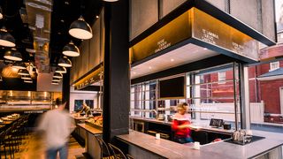 A blurry image of a man walking through a restaurant between dining tables and a bar area. To the left of the bar are floor-to-ceiling windows.