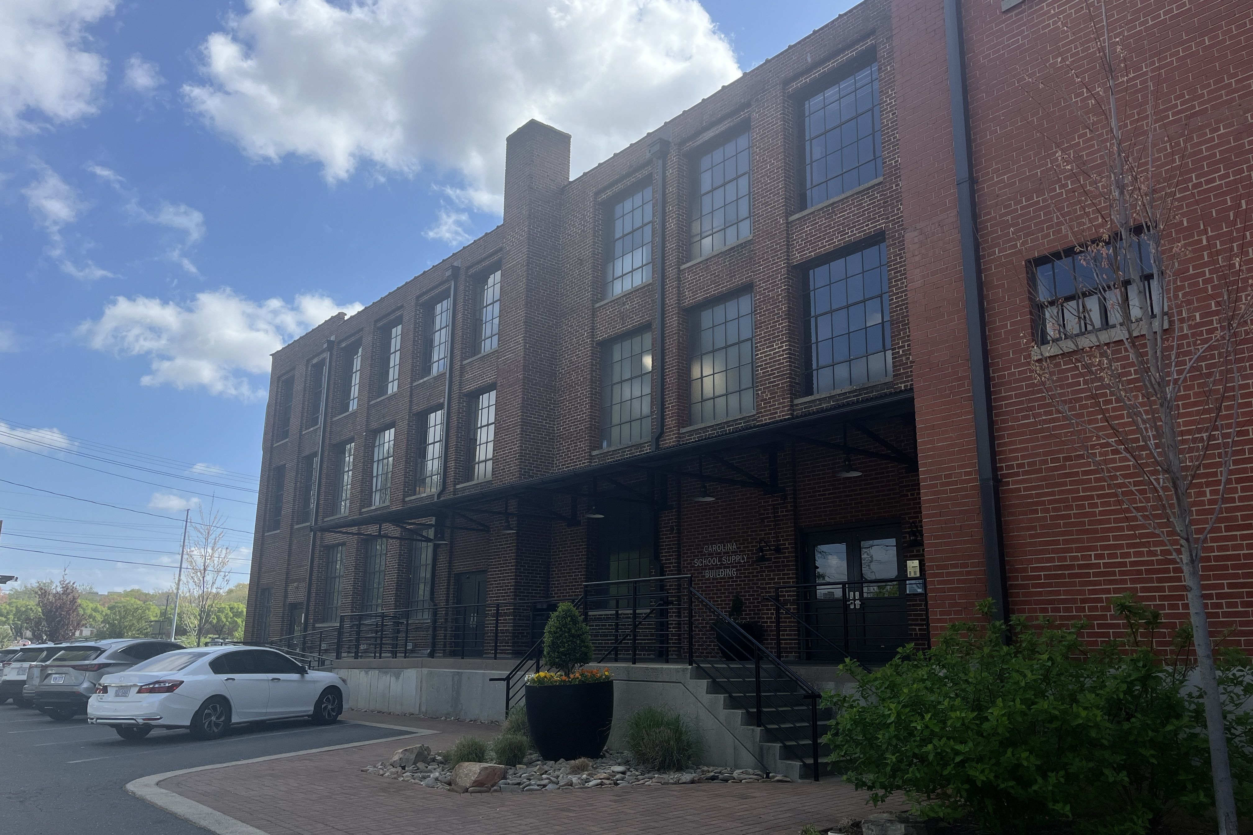 Brick building with large grid windows and a black metal awning over the entrance. Stairs and ramp lead to the door; a parking lot with white cars and a planter of yellow flowers under a blue sky.