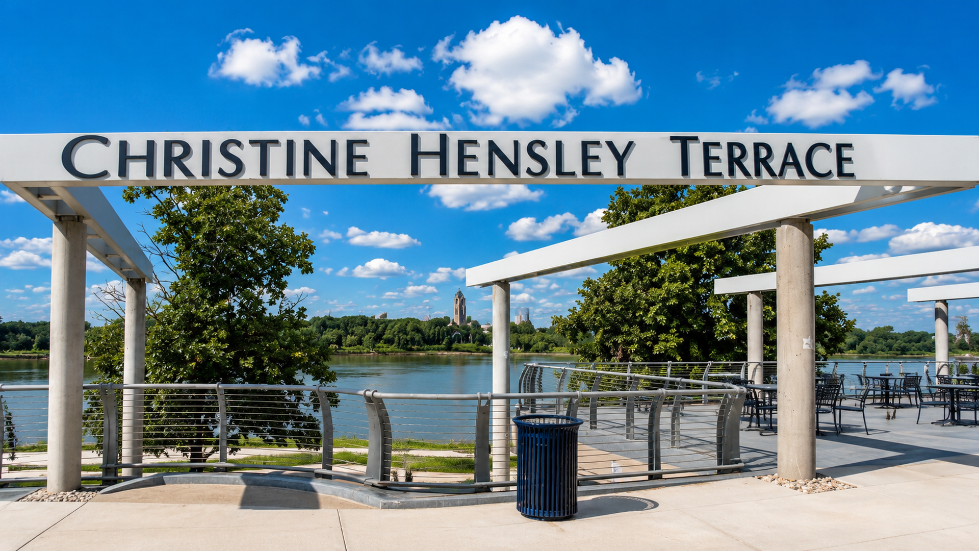 Outdoor terrace with a white sign reading "CHRISTINE HENSLEY TERRACE" across concrete pillars, blue sky with clouds, river and trees, and metal seating along a railing.