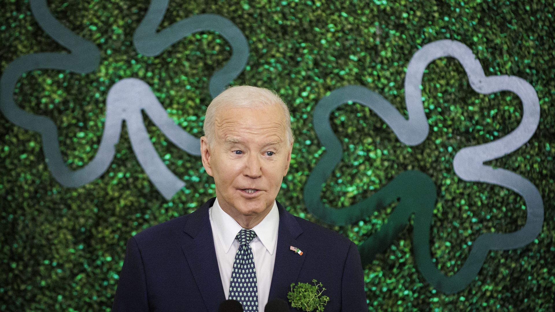 President Joe Biden, wearing a dark blue suit, white shirt and green tie, stands in front fo a sparkly green backdrop with two four-leaf clovers.