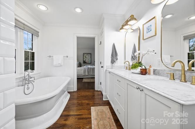 Bright bathroom with white walls, a clawfoot bathtub, double sink with gold fixtures, large mirror, wooden floor, and doorway leading to a bedroom. Natural light enters through two windows.