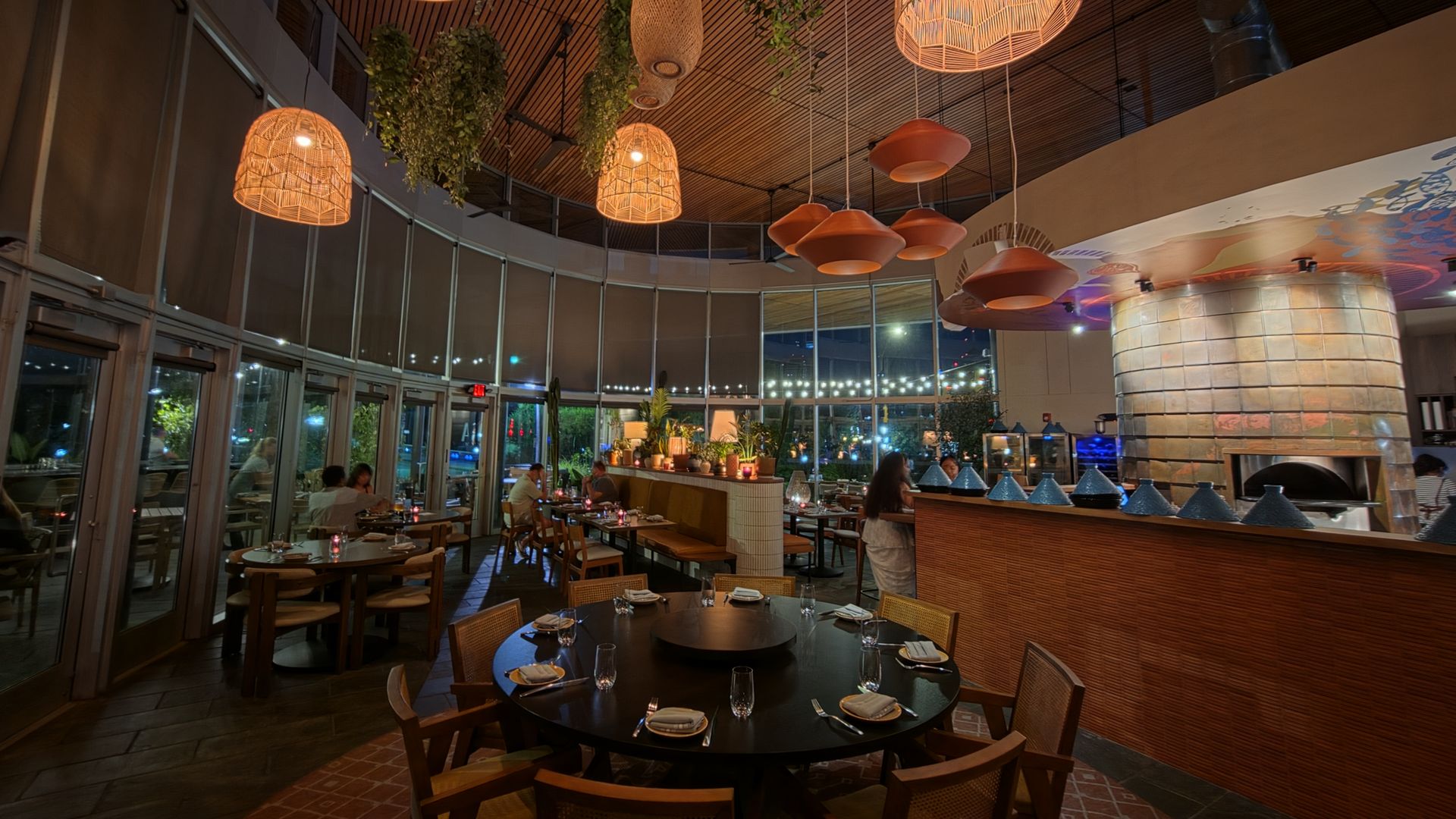 Interior of a stylish restaurant with warm orange wicker pendant lamps, hanging greenery, a wooden slat ceiling, and round tables set for dinner by glass walls.
