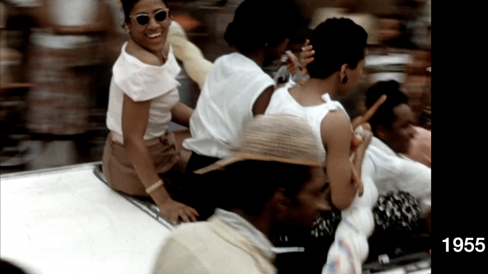 Woman in a white shirt and sunglasses sitting in the back of a convertible looking back.