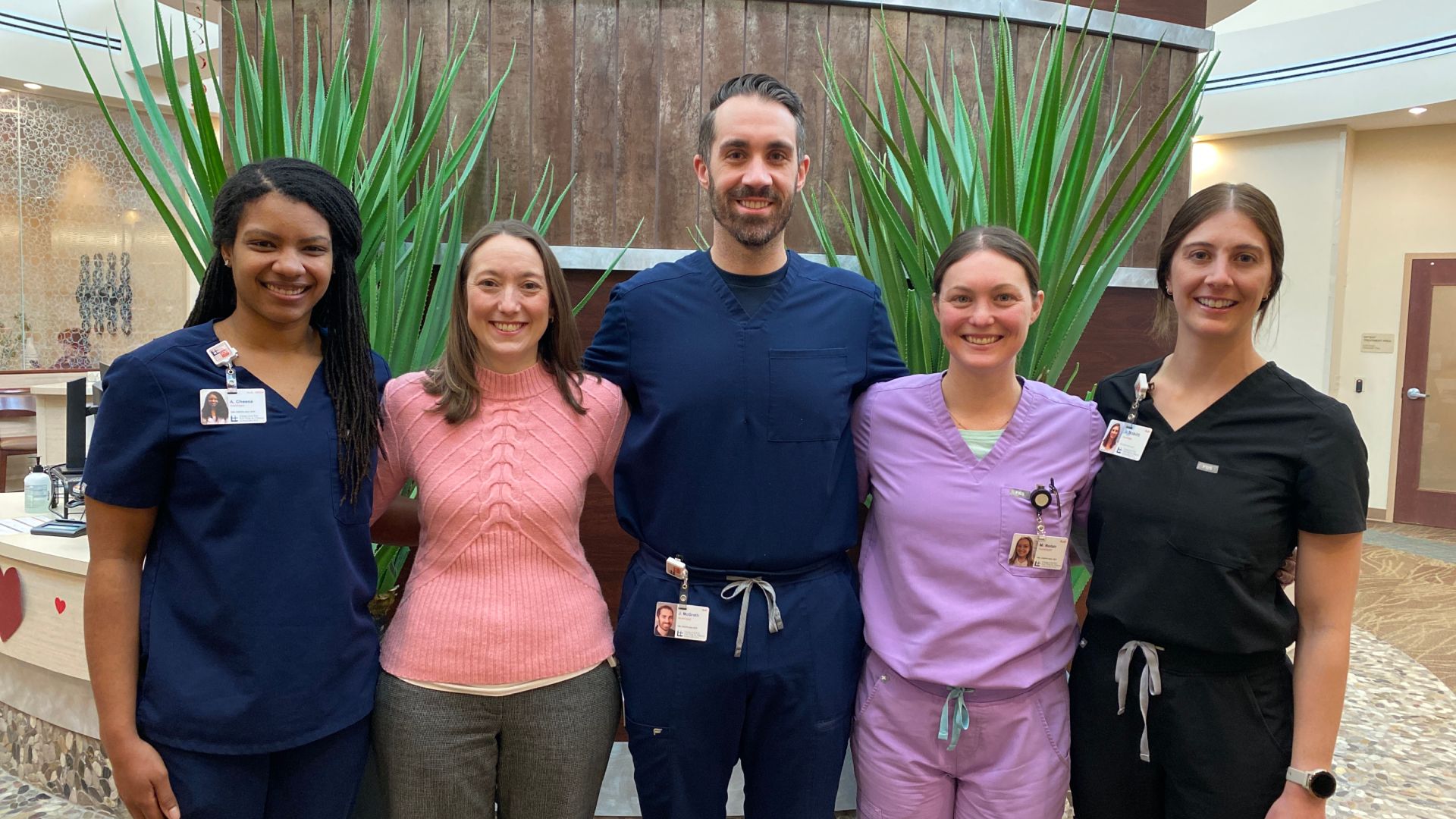 Five health care workers stand in a hospital lobby with tall green plants behind them; they wear navy scrubs, pink sweater, lavender scrubs and black scrubs, all smiling with ID badges.