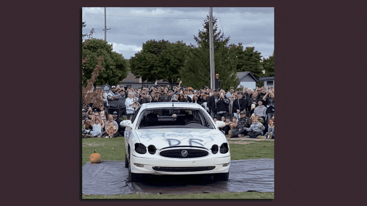 A giant pumpkin falling onto an old white car with its headlights removed and graffiti on the hood, as a crowd watches.