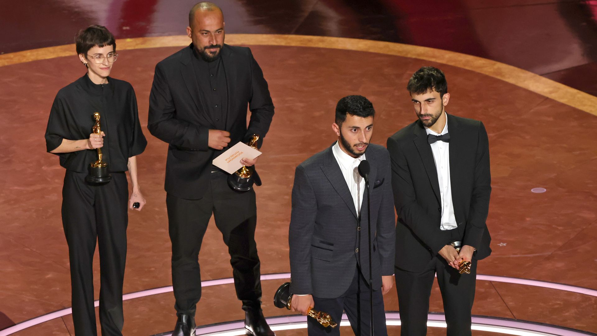 Four men on the oscars stage, while one is speaking while holding the award. 