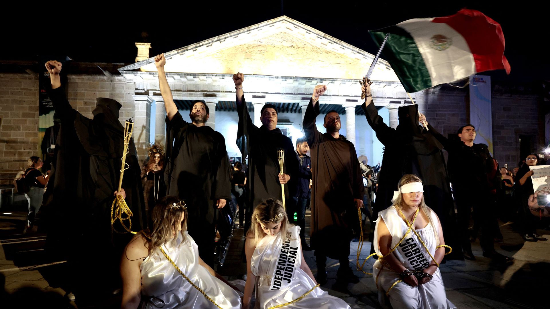 Four men in black robes hold their fists in the air while protesting. One is holding a large Mexican flag. In front of the men, three women dressed in white robes and sashes with messages sit on their knees