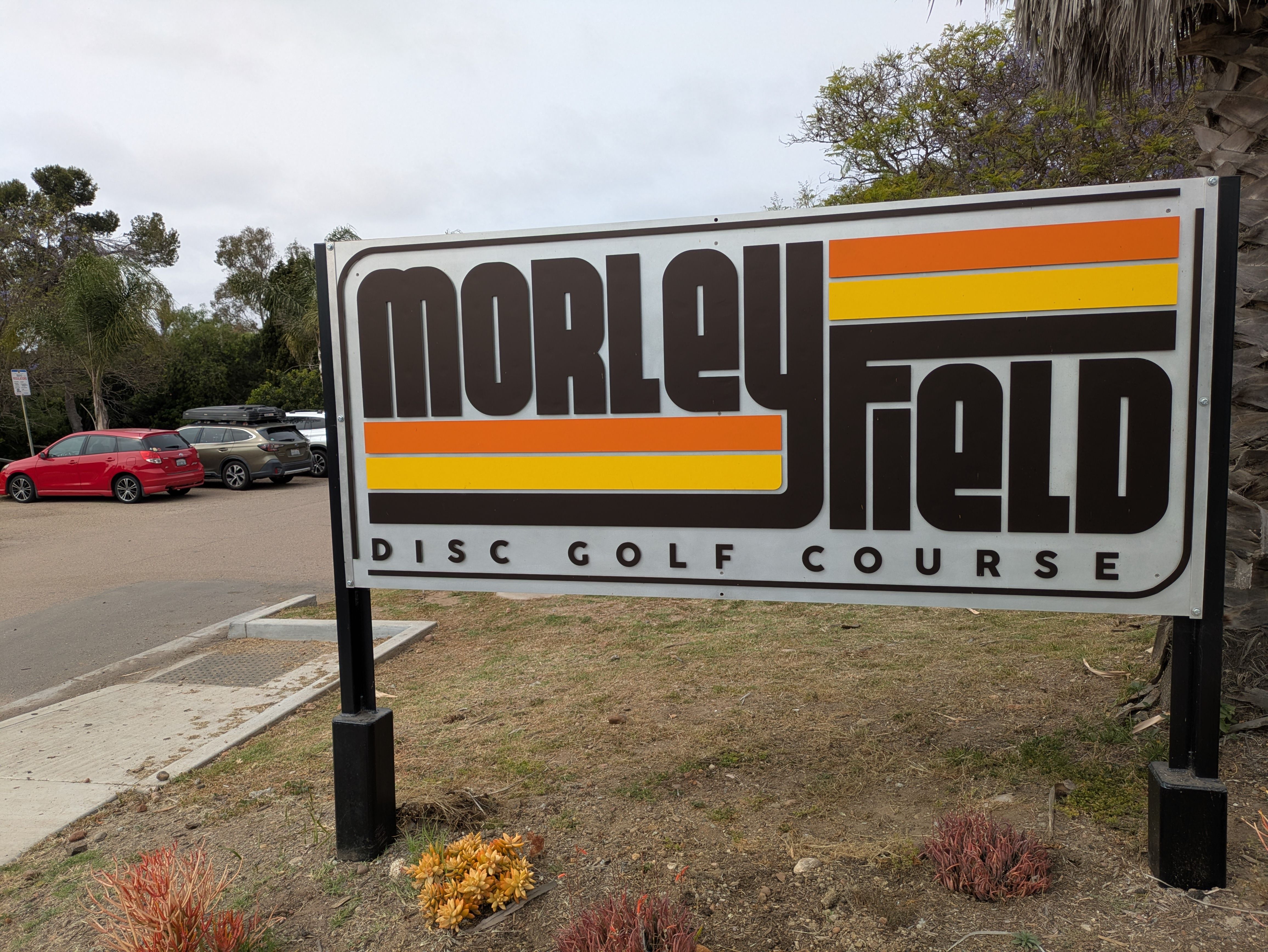 Sign reads "Morley Field Disc Golf Course" in bold black with orange and yellow stripes; behind are parked cars and trees on a cloudy day.