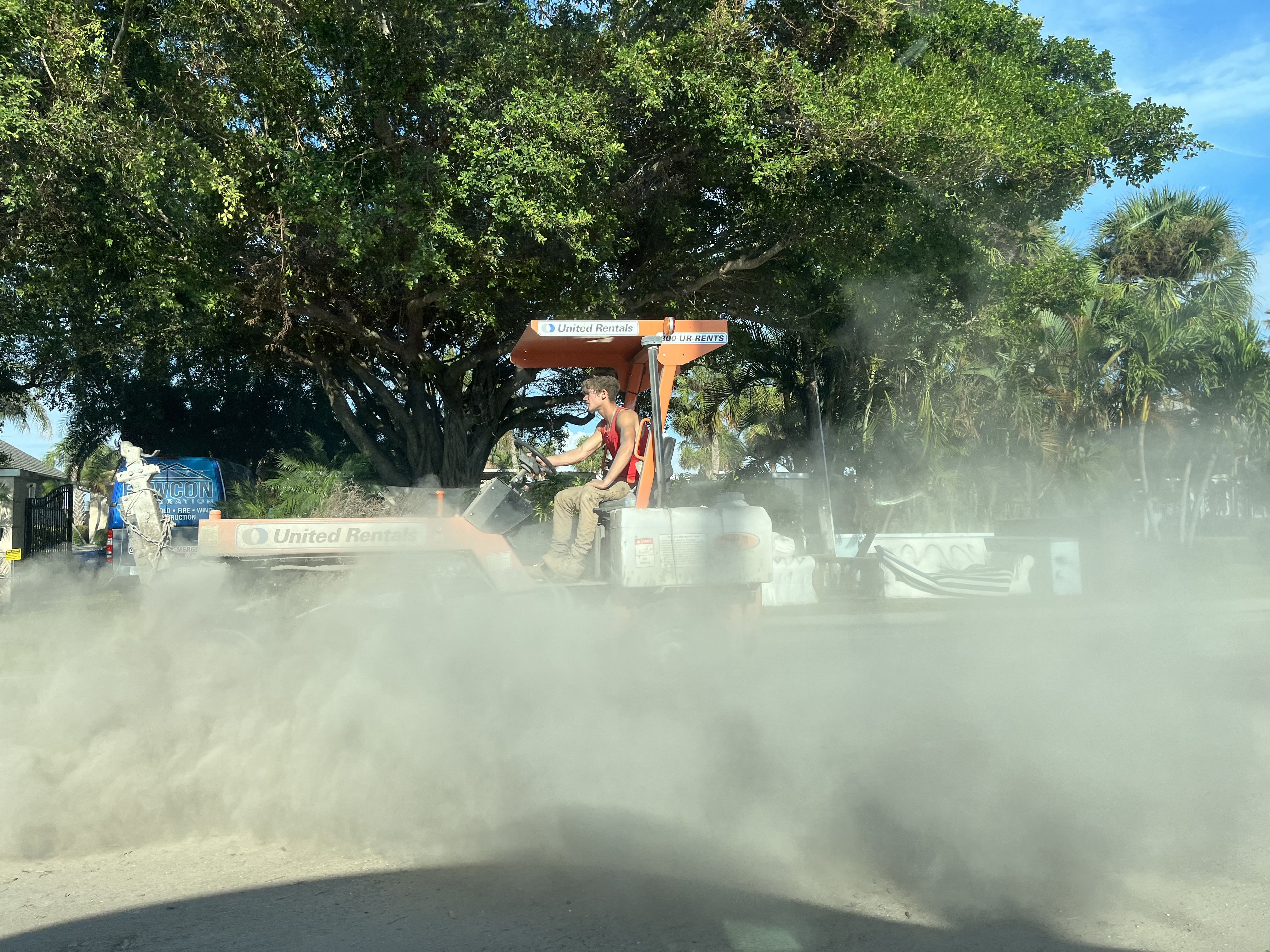 A man drives an orange street sweeper surrounded by a plume of sand.