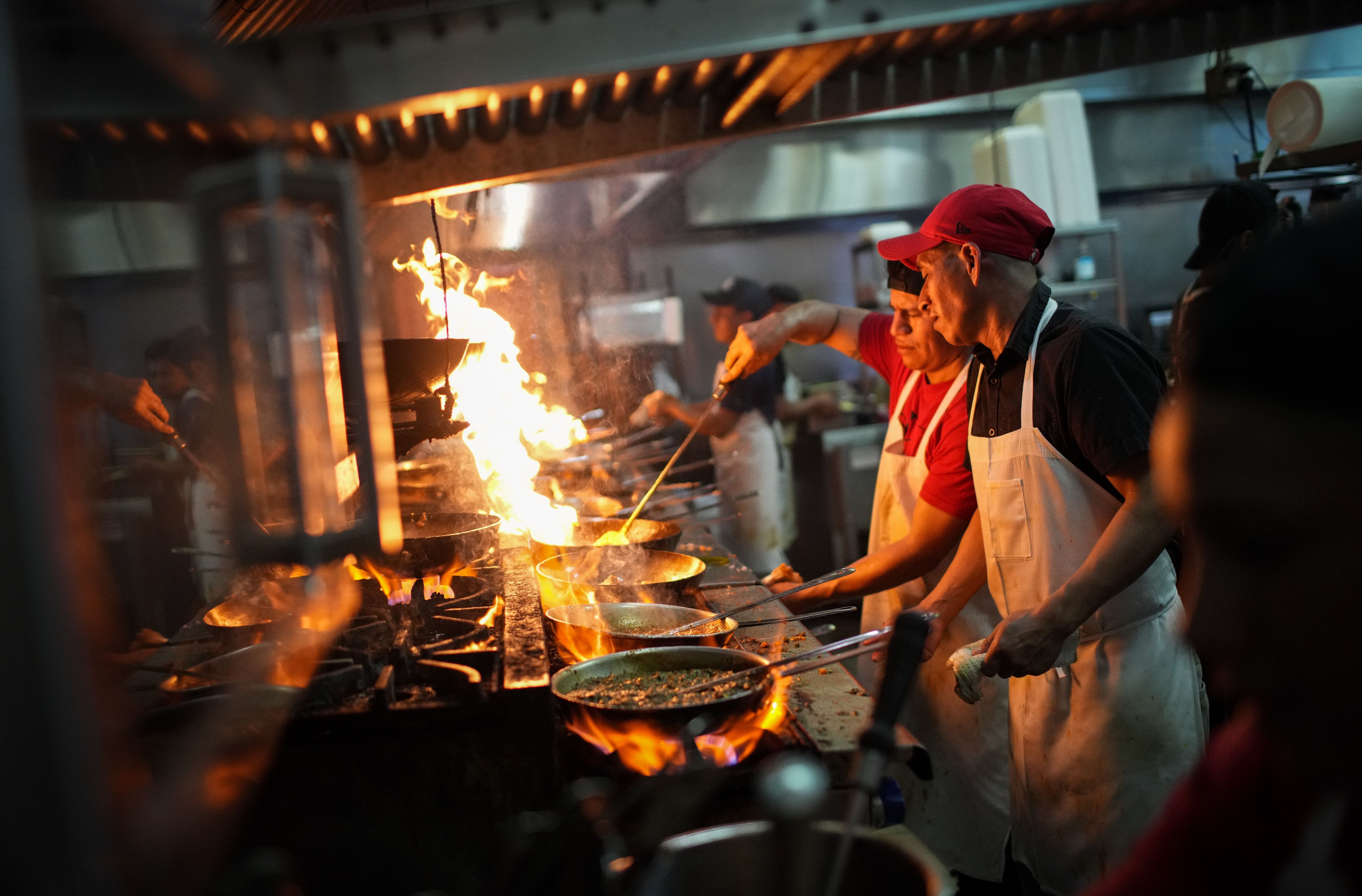 Two people work at a stove in a restaurant kitchen, handing a saucepan as flames rise up