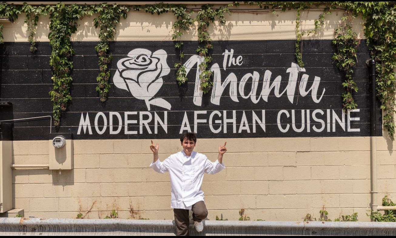 Man in white chef coat posing with raised hands in front of a black and beige wall mural with green ivy. The mural reads "the Mantu MODERN AFGHAN CUISINE" with a white rose graphic.