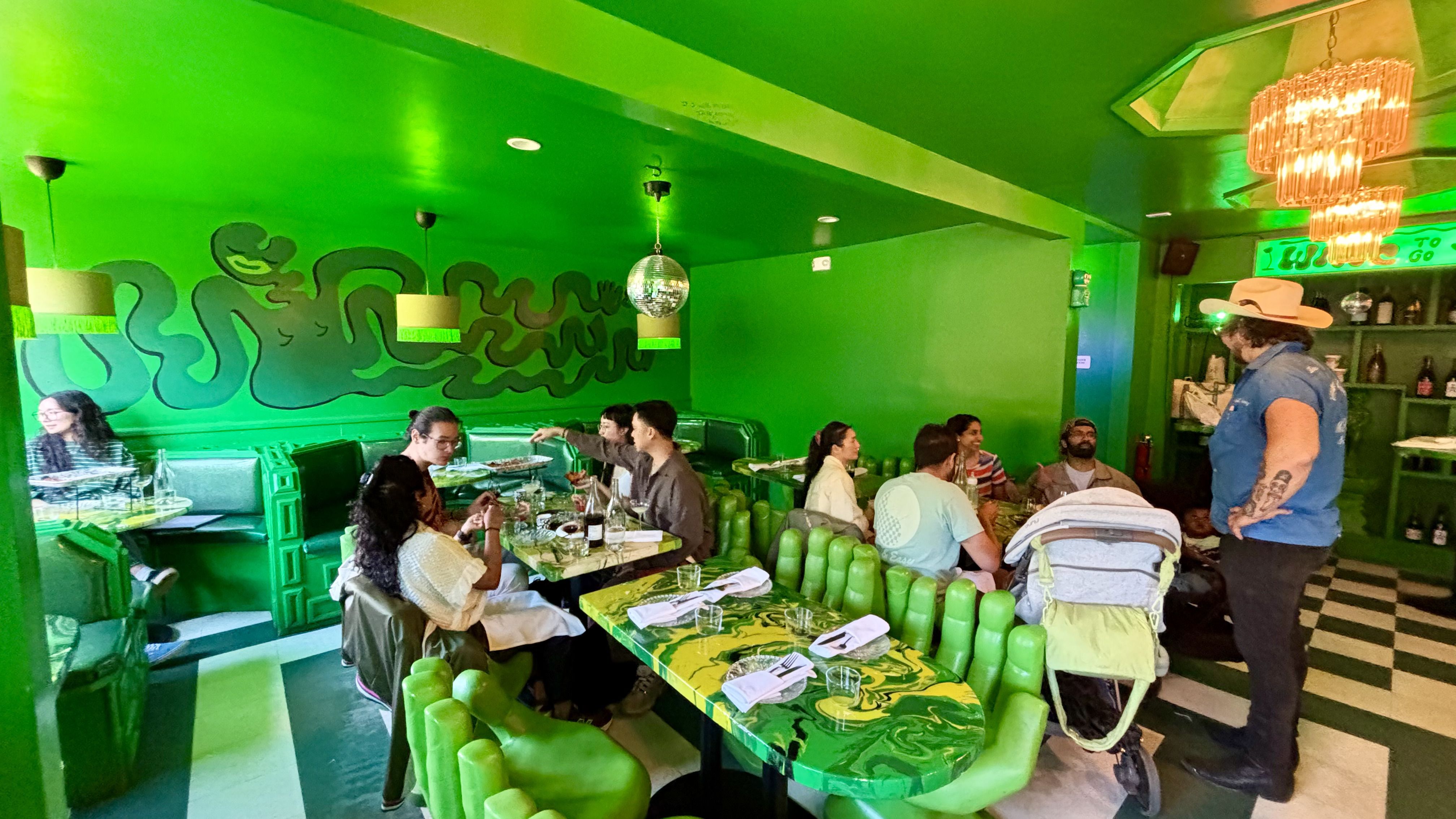 Bright green-themed restaurant interior with green walls, green chairs, and a green ceiling. A group of people are seated at tables with green marble patterns, chatting and dining.