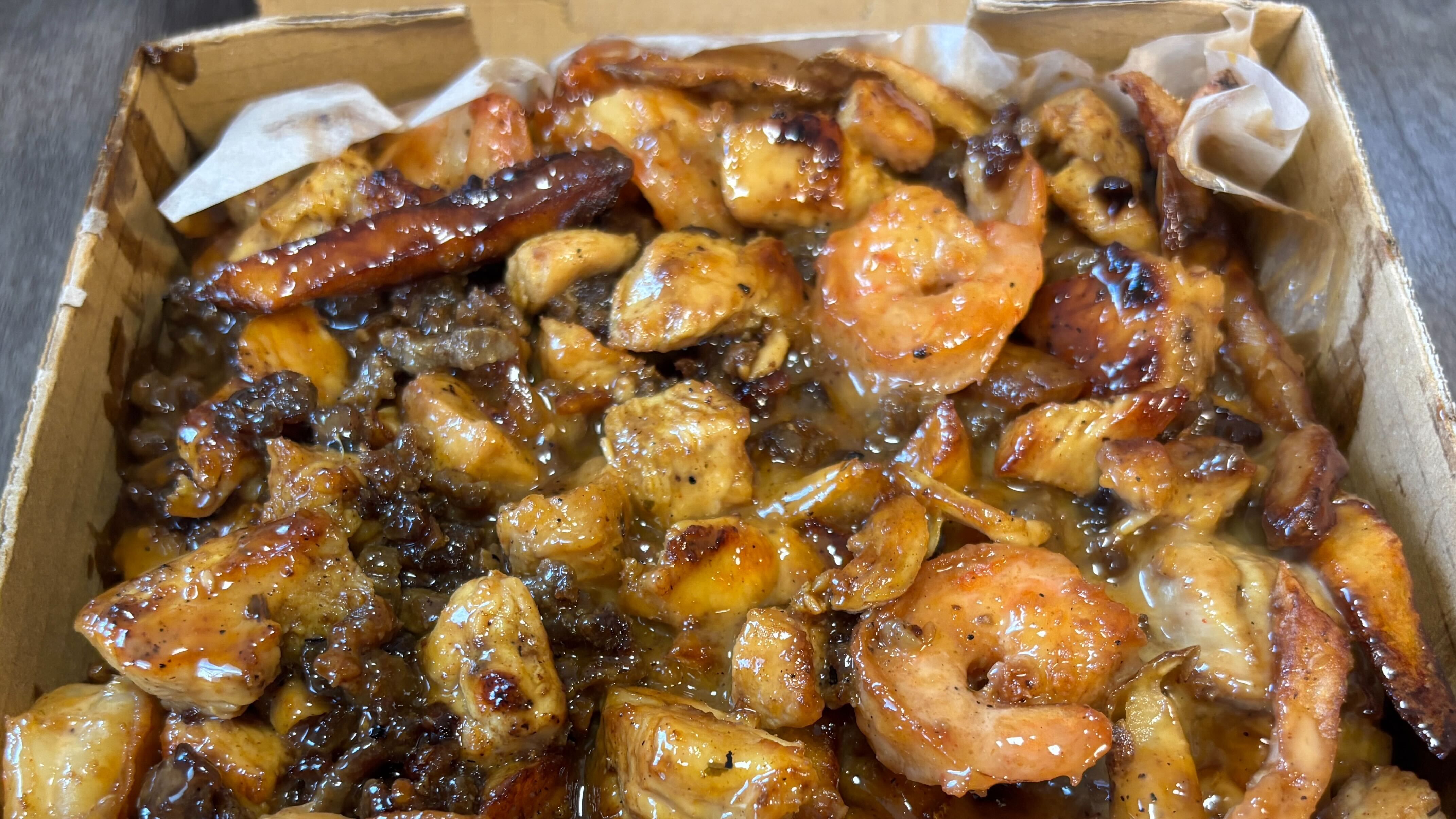Close-up of a cardboard container filled with glazed grilled shrimp, chunks of chicken, and pieces of grilled meat, all coated in a shiny brown sauce.