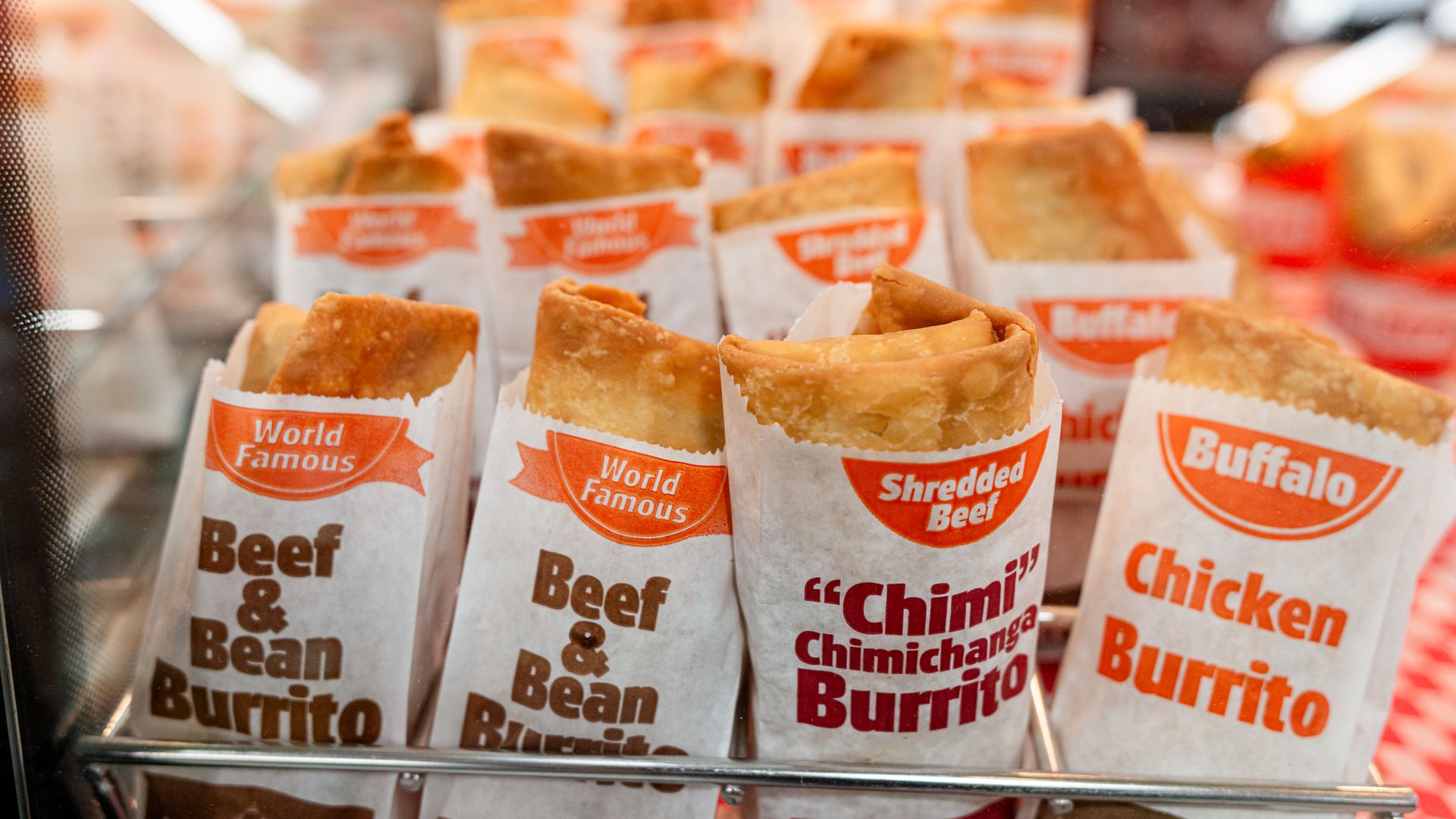 Row of warm burritos wrapped in white paper with orange 'World Famous' logos, displayed behind glass; labels read 'Beef & Bean Burrito' and 'Chimichanga Burrito'.