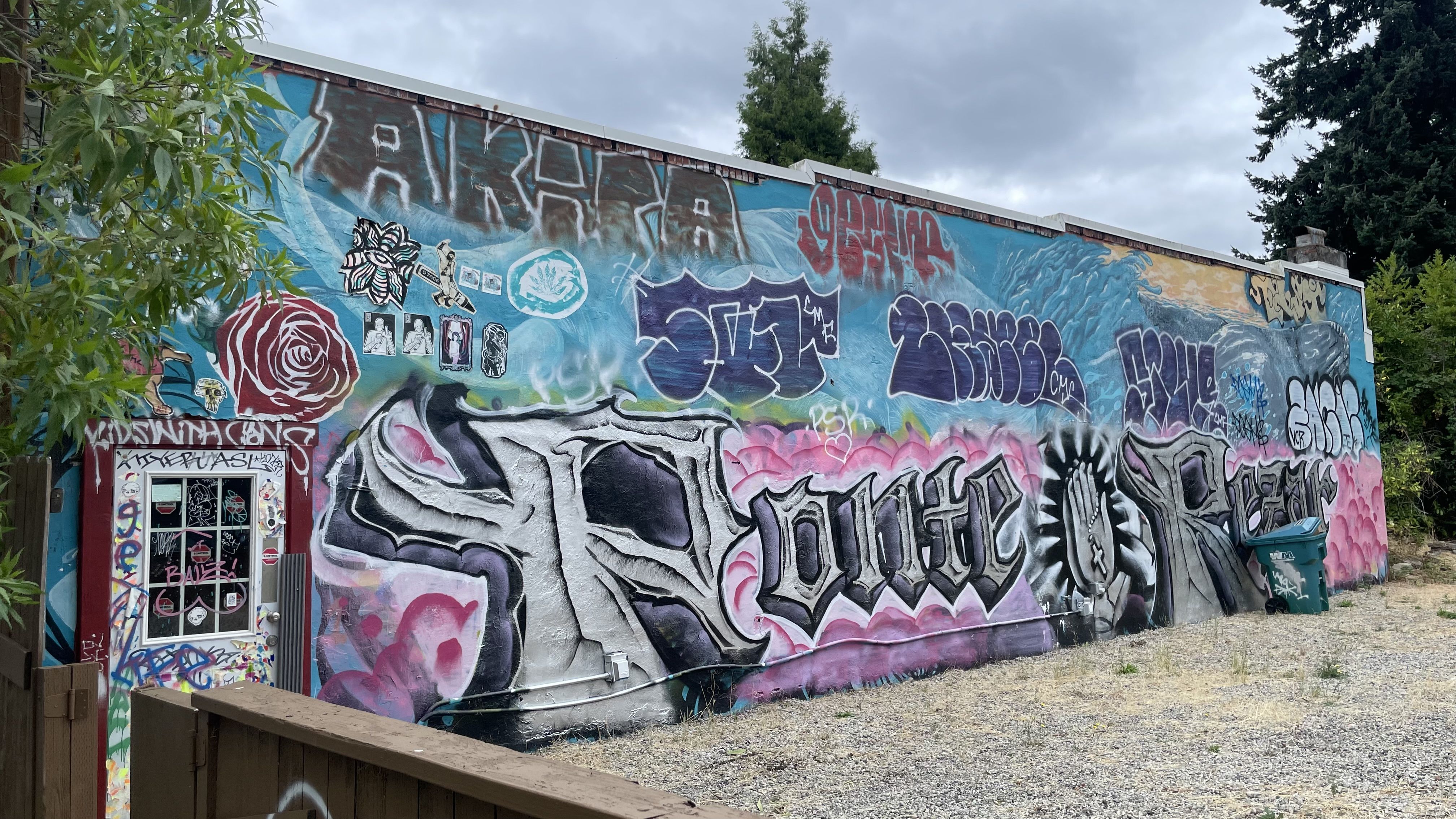Colorful graffiti mural on a building wall featuring large stylized letters, a red rose, praying hands, and various tags under a cloudy sky with trees nearby.