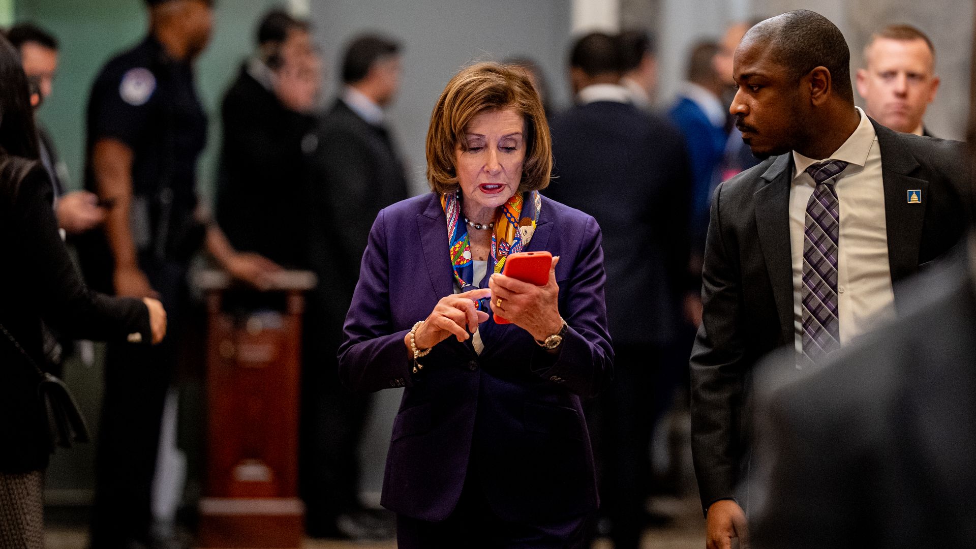 House Speaker Nancy Pelosi, wearing a purple suit and typing on her phone surrounded by her security detail.