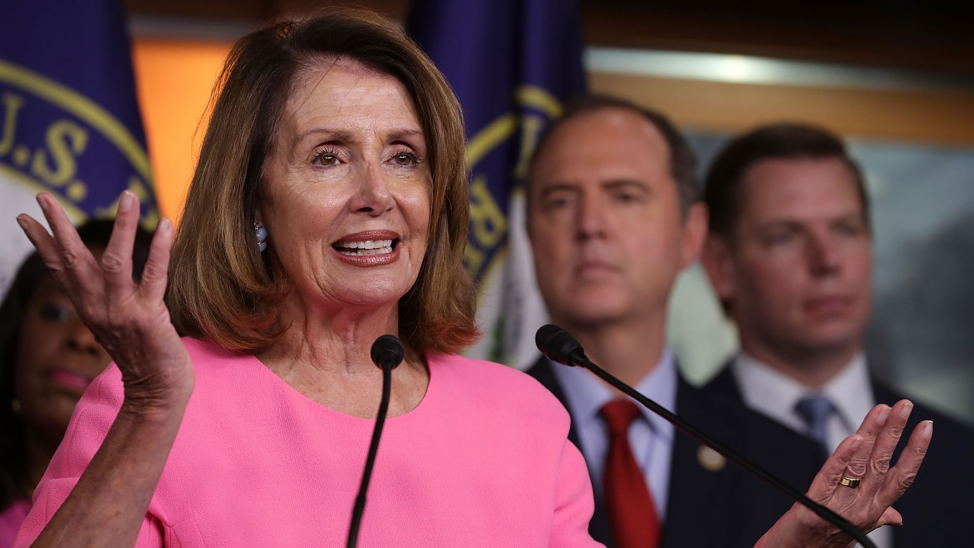 Nancy Pelosi in pink standing in front of Democrats raises her arms in a shrug smiling.