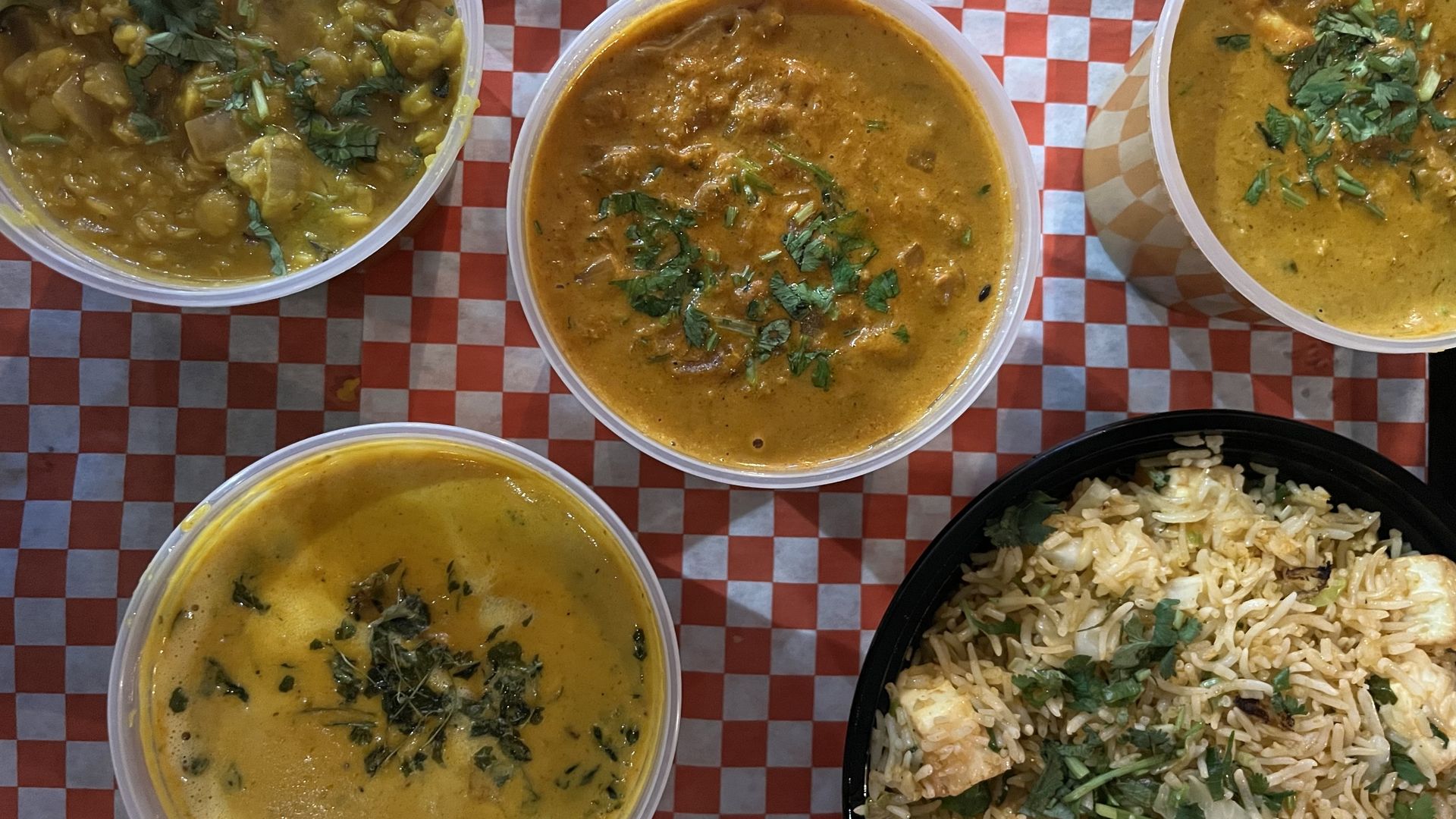 Top-down view of four plastic bowls of Indian curry (yellow and orange) garnished with cilantro on a red-and-white checkered surface, beside a black bowl of seasoned rice.