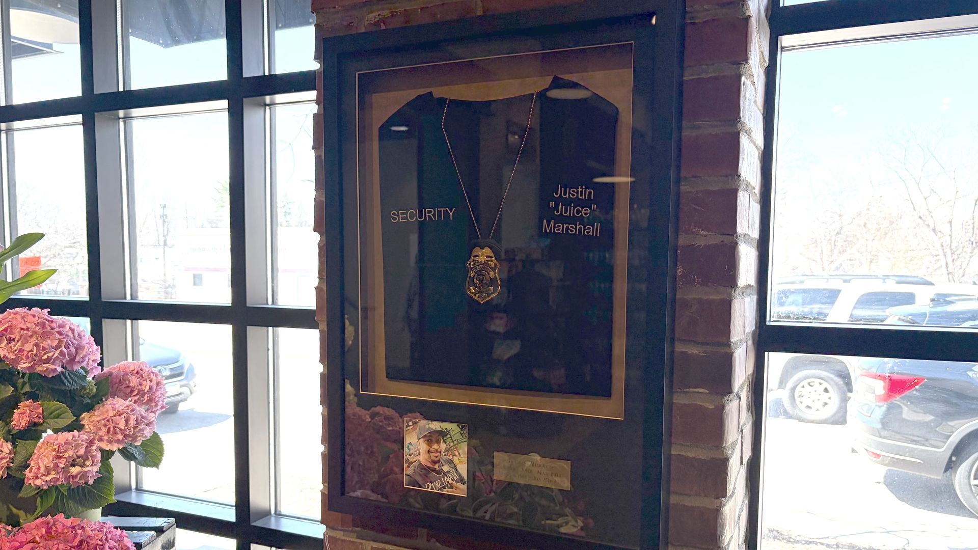 Framed display on a brick pillar features a security badge necklace on a dark shirt; labels read "SECURITY" and "Justin \"Juice\" Marshall". Pink hydrangeas sit to the left, with large windows and parked cars outside.