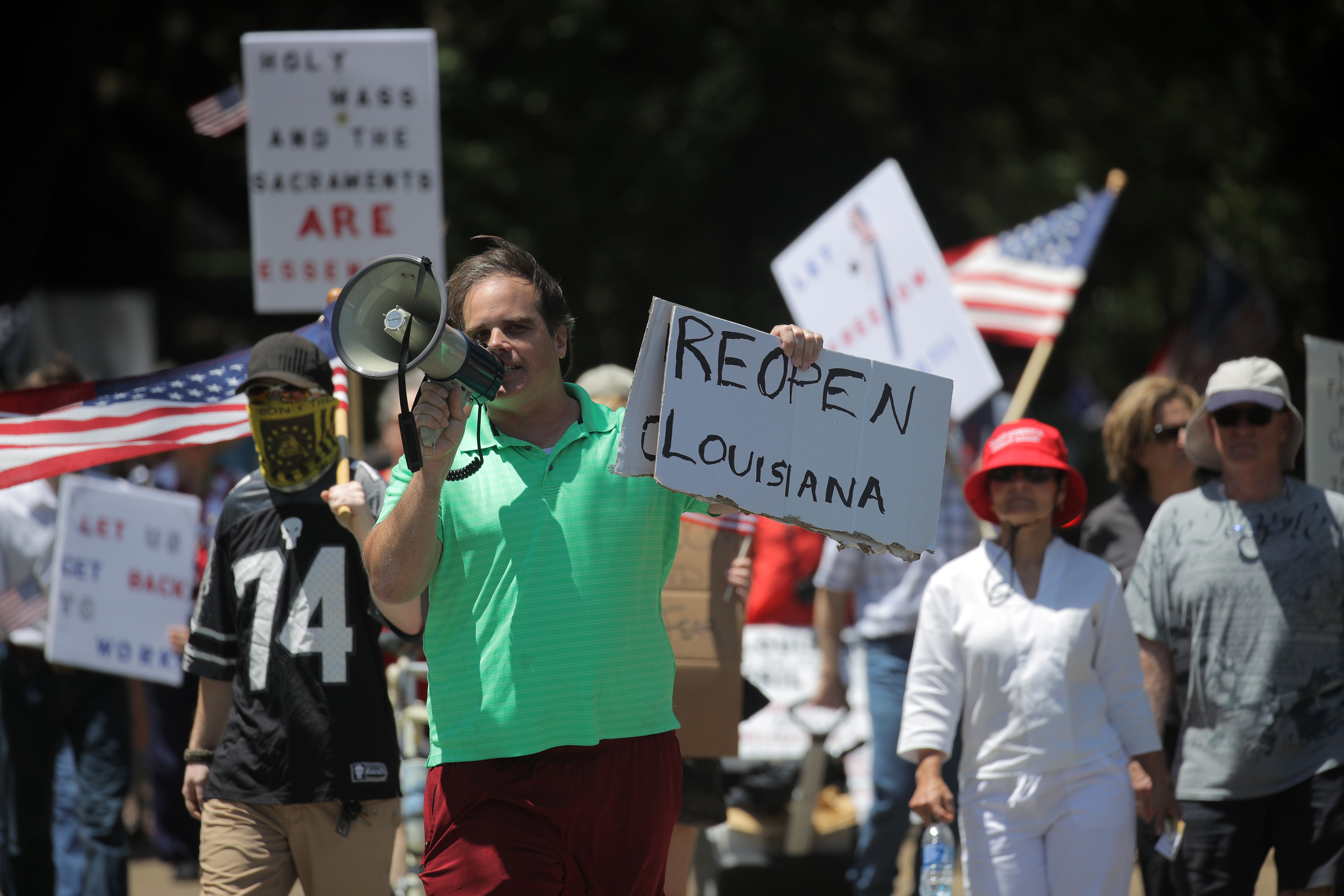In this image, a man holds a cardboard sign that reads "reopen louisiana" and a megaphone