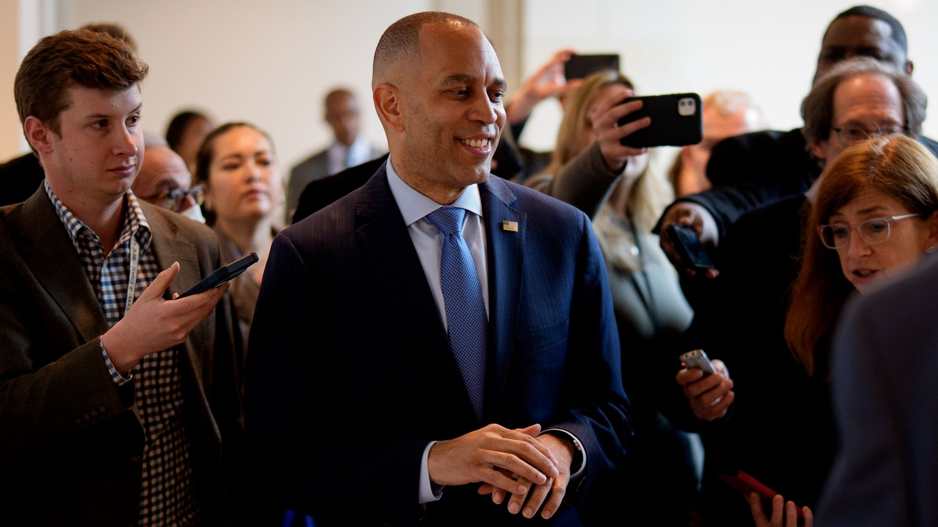 House Minority Leader Hakeem Jeffries wearing a blue suit and smiling while surrounded by a throng of reporters in a white hallway.