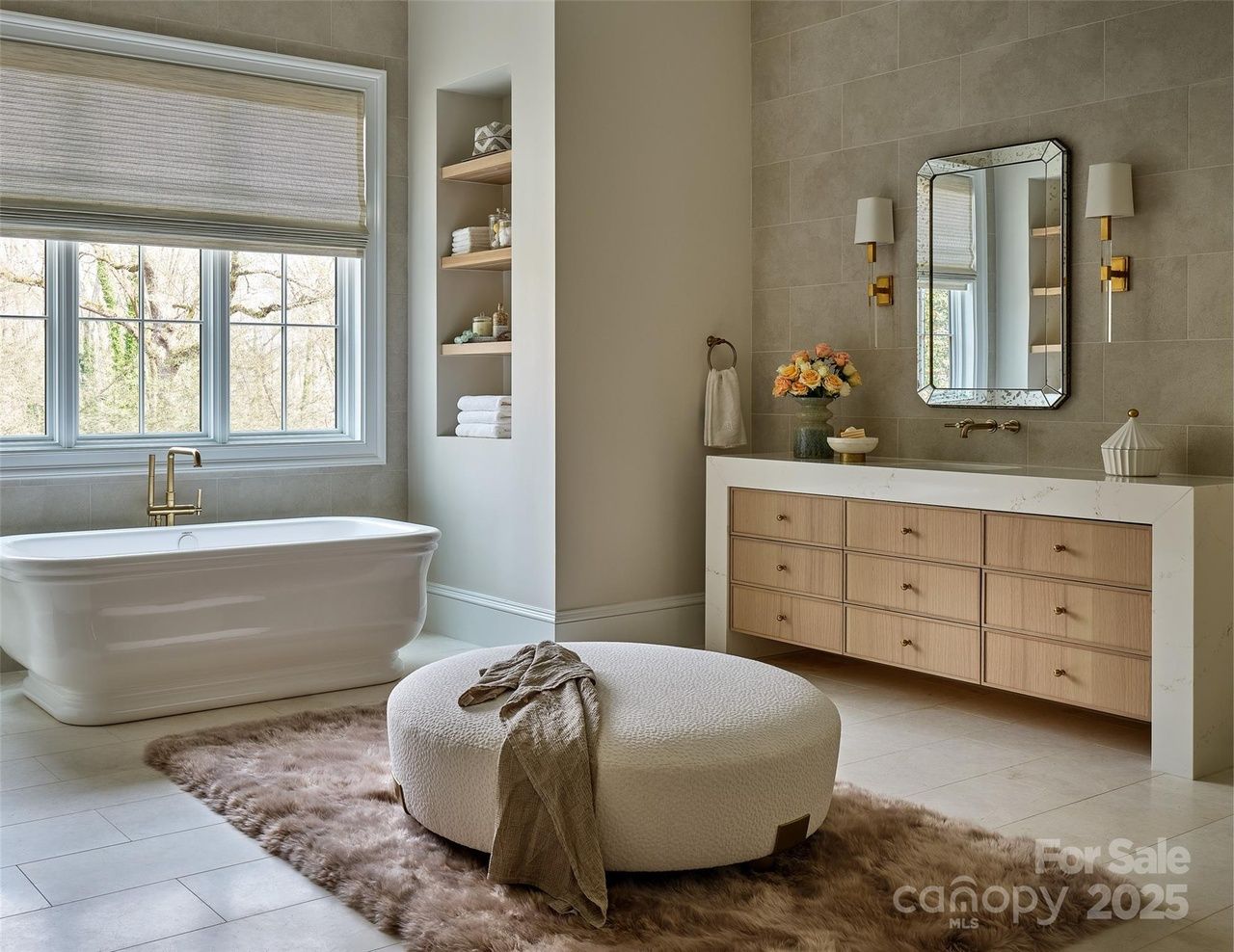 Bright bathroom with white freestanding tub, large window, wooden vanity with marble countertop, decorative mirror, flowers, and circular ottoman on brown plush rug.