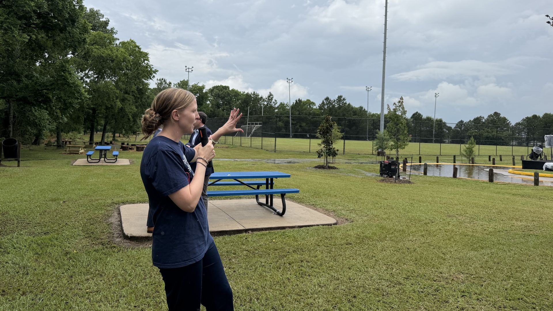 Two people in a grassy park: a girl in a navy shirt raises her hand while others watch. In the background there are picnic tables, a fence, and a tall light pole under a cloudy sky.