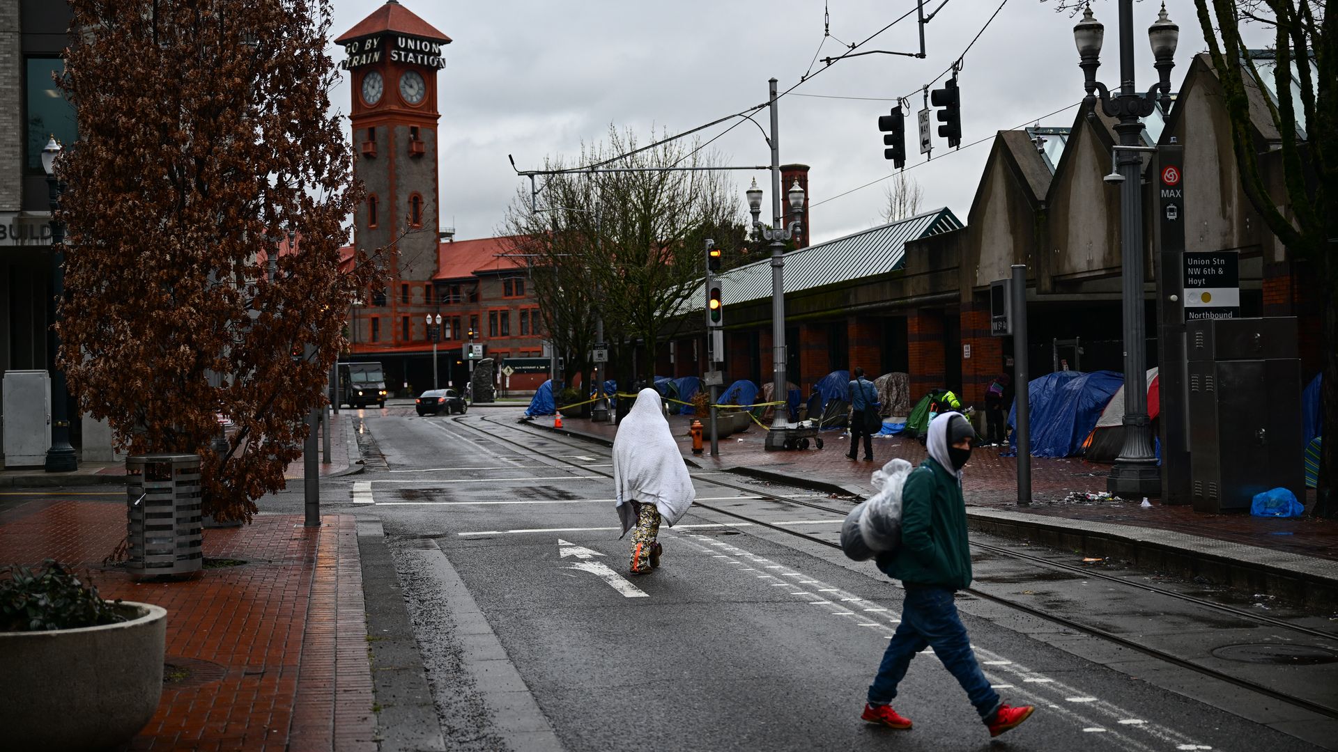 A photo of a person walking with a blanket covering them in front of a sidewalk lined with tents.