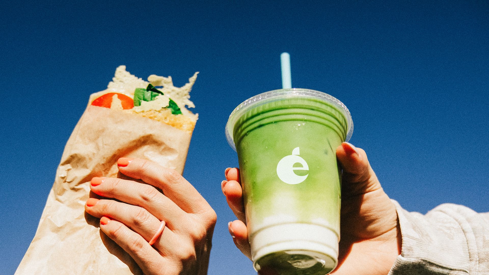 Two hands hold food items against a clear blue sky: a sandwich wrapped in brown paper with visible tomato and lettuce, and a green and white iced drink in a plastic cup with a straw.