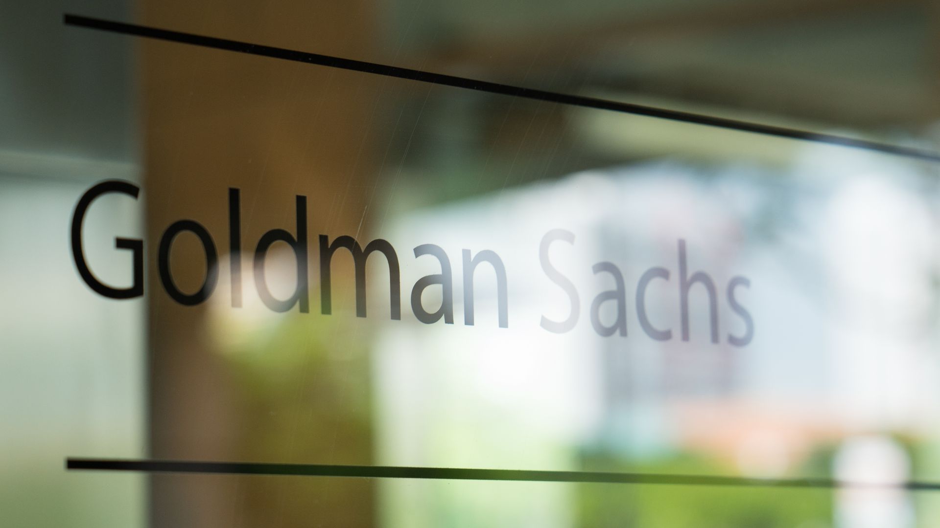 Close-up of a glass sign with the partially visible text "Goldman Sachs" in bold black letters, a diagonal black line across the pane, and a blurred green background.