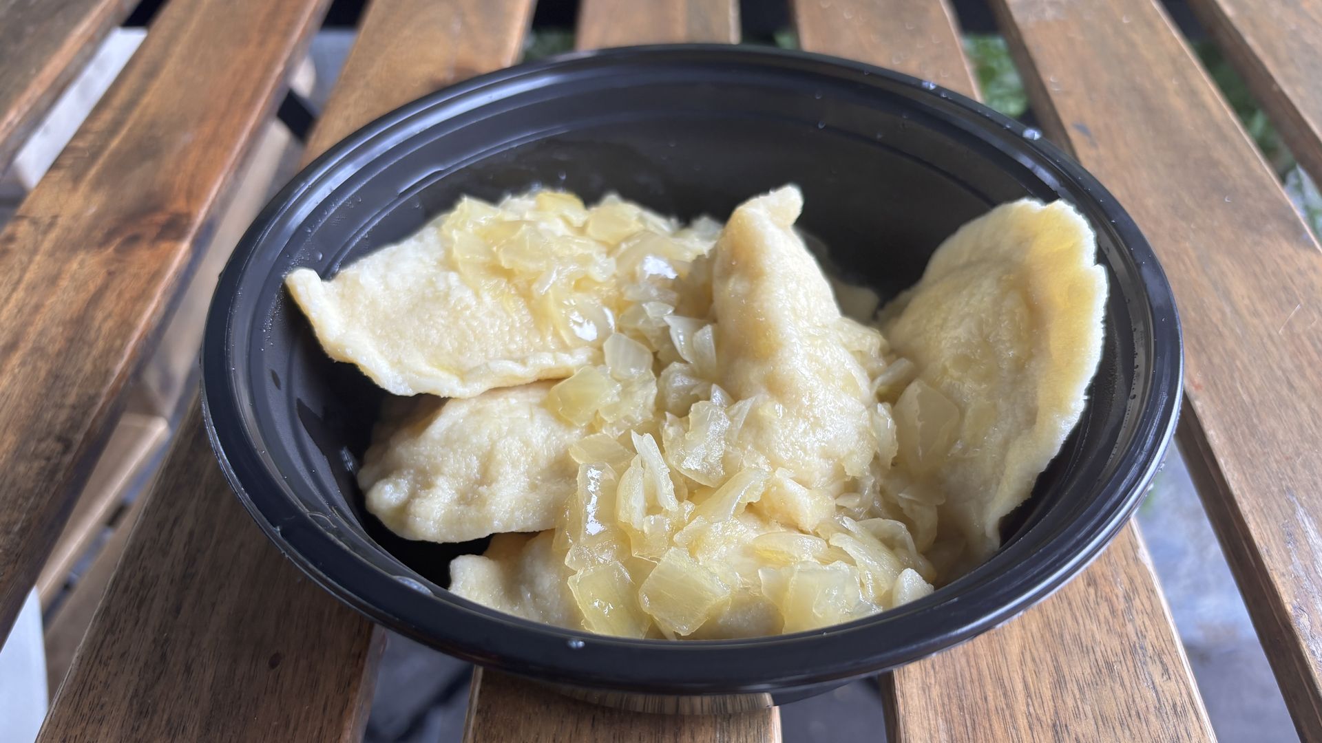 Black bowl on wooden table with several pierogi dumplings topped with cooked, diced onions.