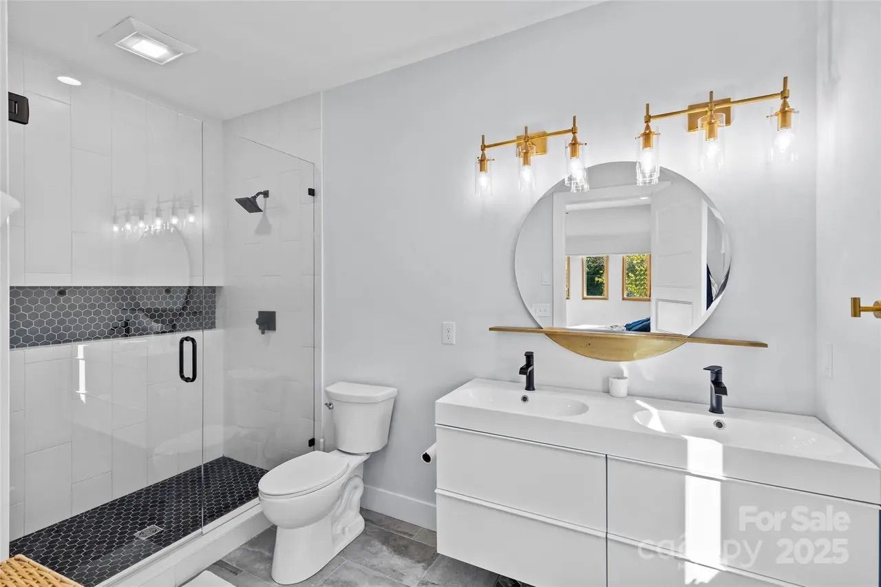 Modern bathroom with white walls, double sink vanity, round mirror with gold shelf, gold light fixtures, glass shower with black hex tile floor and accent wall, white toilet, and gray floor tiles.